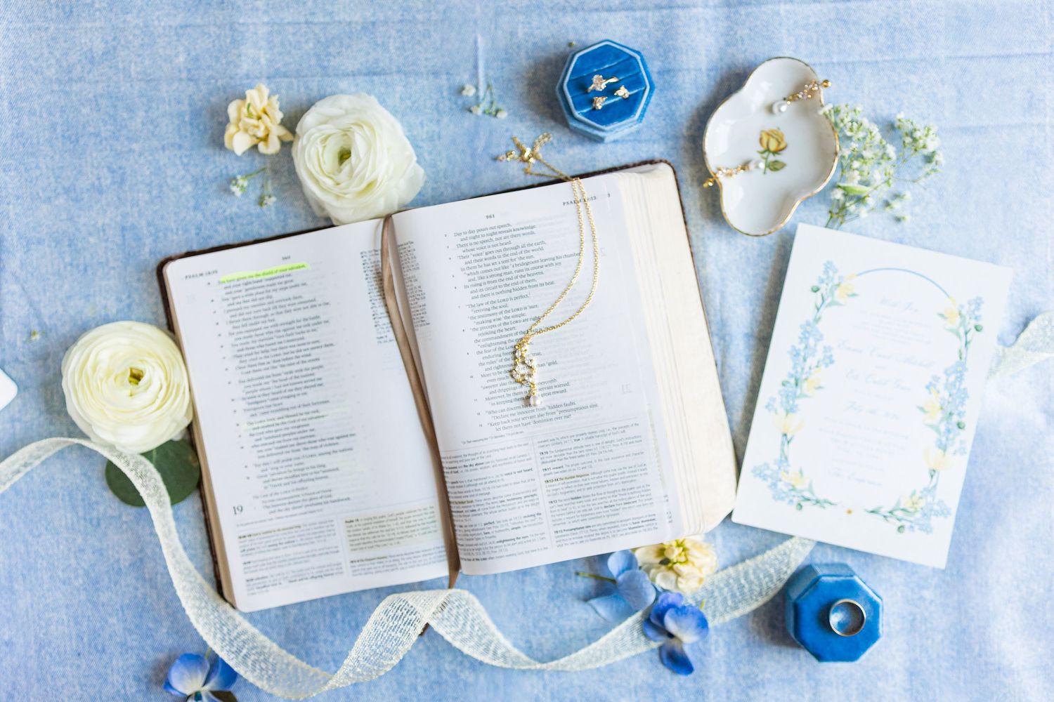 Open Bible with gold necklace, white ranunculus flowers, blue candles, and elegant stationery on light blue linen.