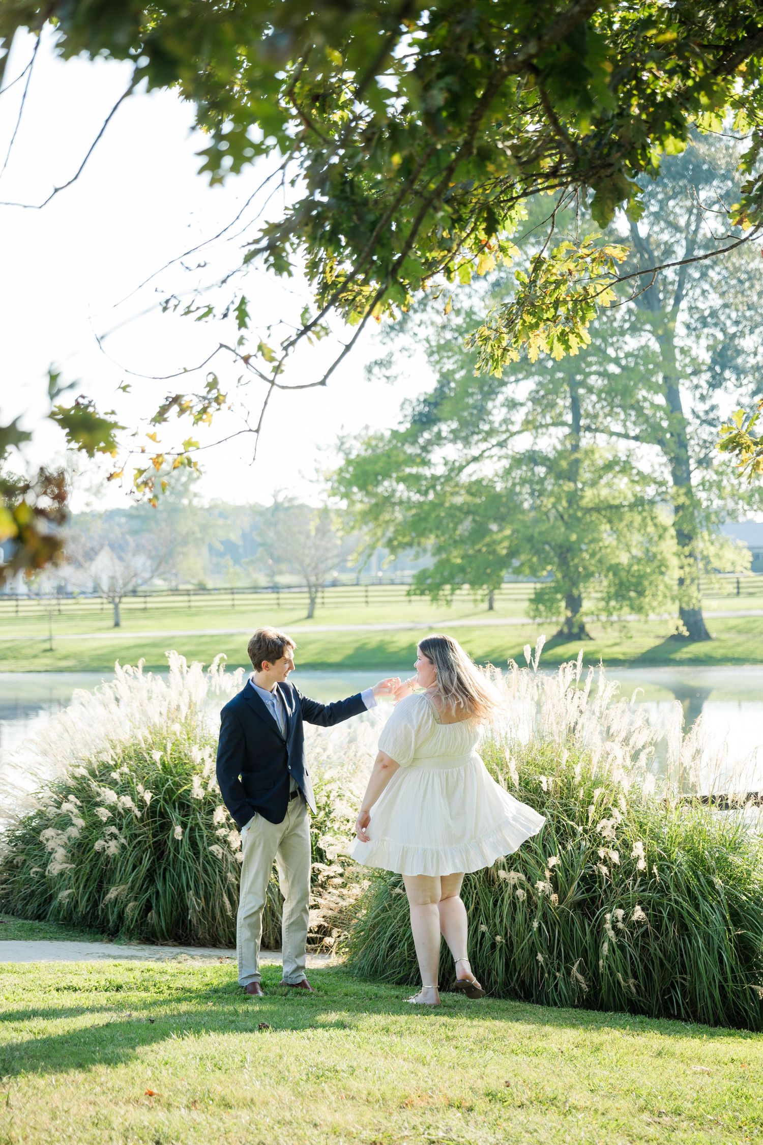 Couple dancing on green lawn near ornamental grasses and pond, man in blazer, woman in white dress.