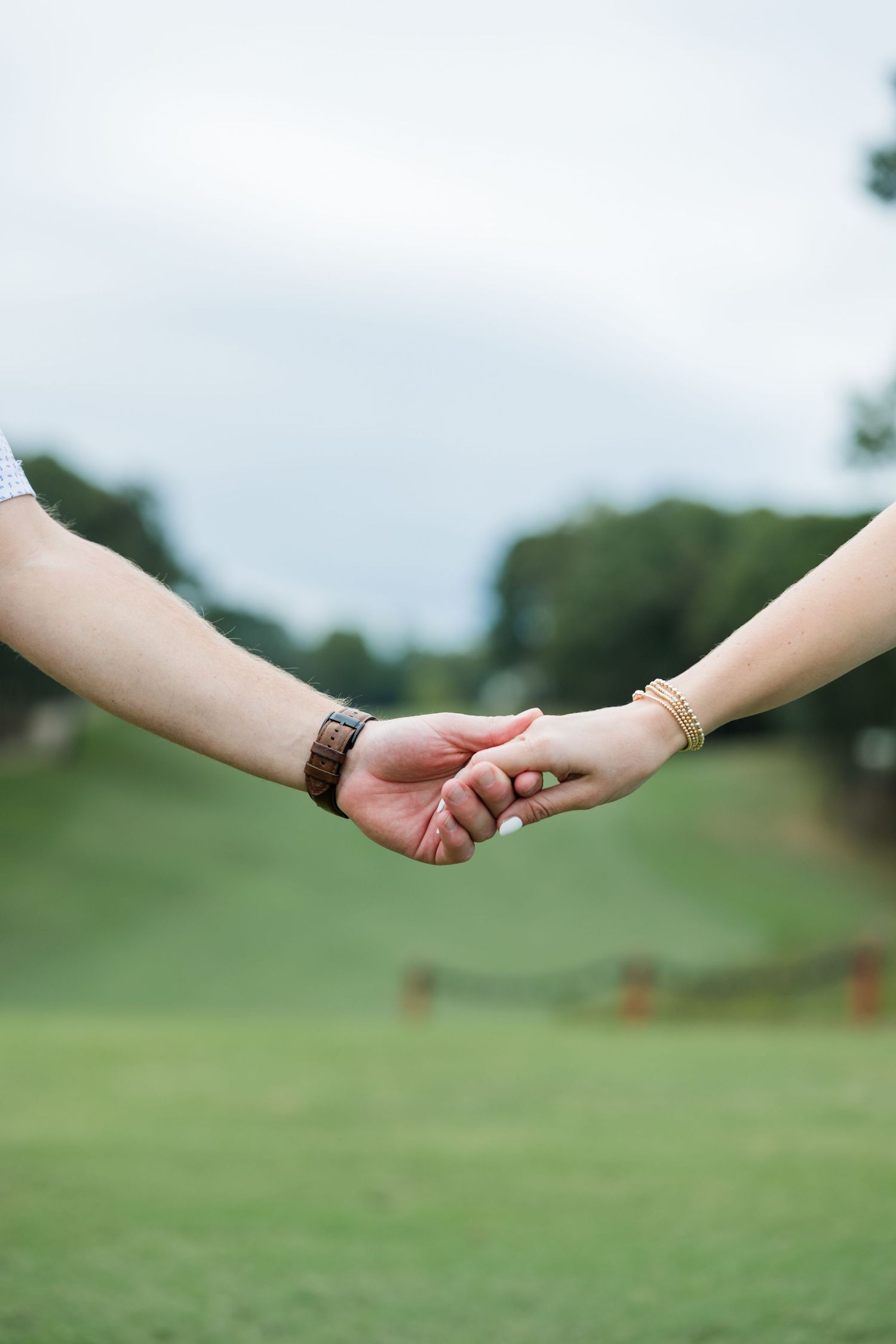 Close-up of a man and woman holding hands outdoors, with a blurred green grassy hillside and trees in the background.
