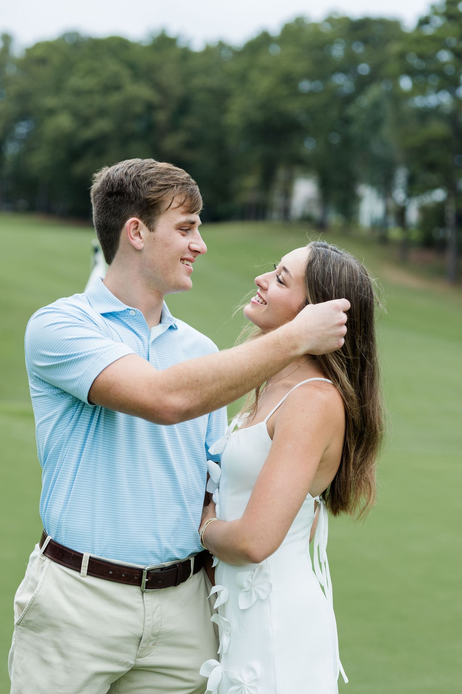 Smiling man in a light blue polo gently tucks hair behind his fiancée's ear on a golf course lawn during an engagement shoot.