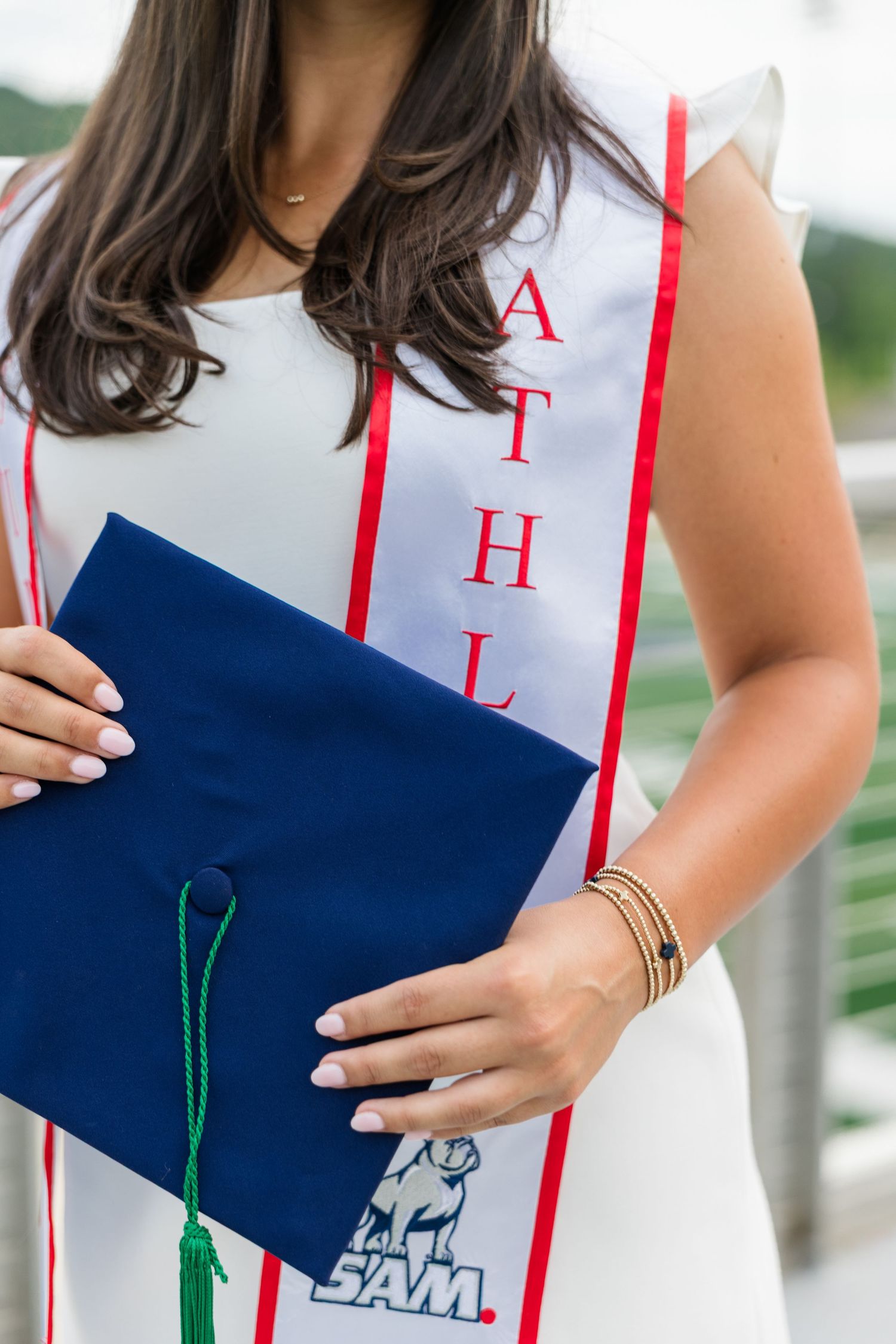 Graduate in white dress holds navy cap with green tassel, wearing SAM university athletic sash.