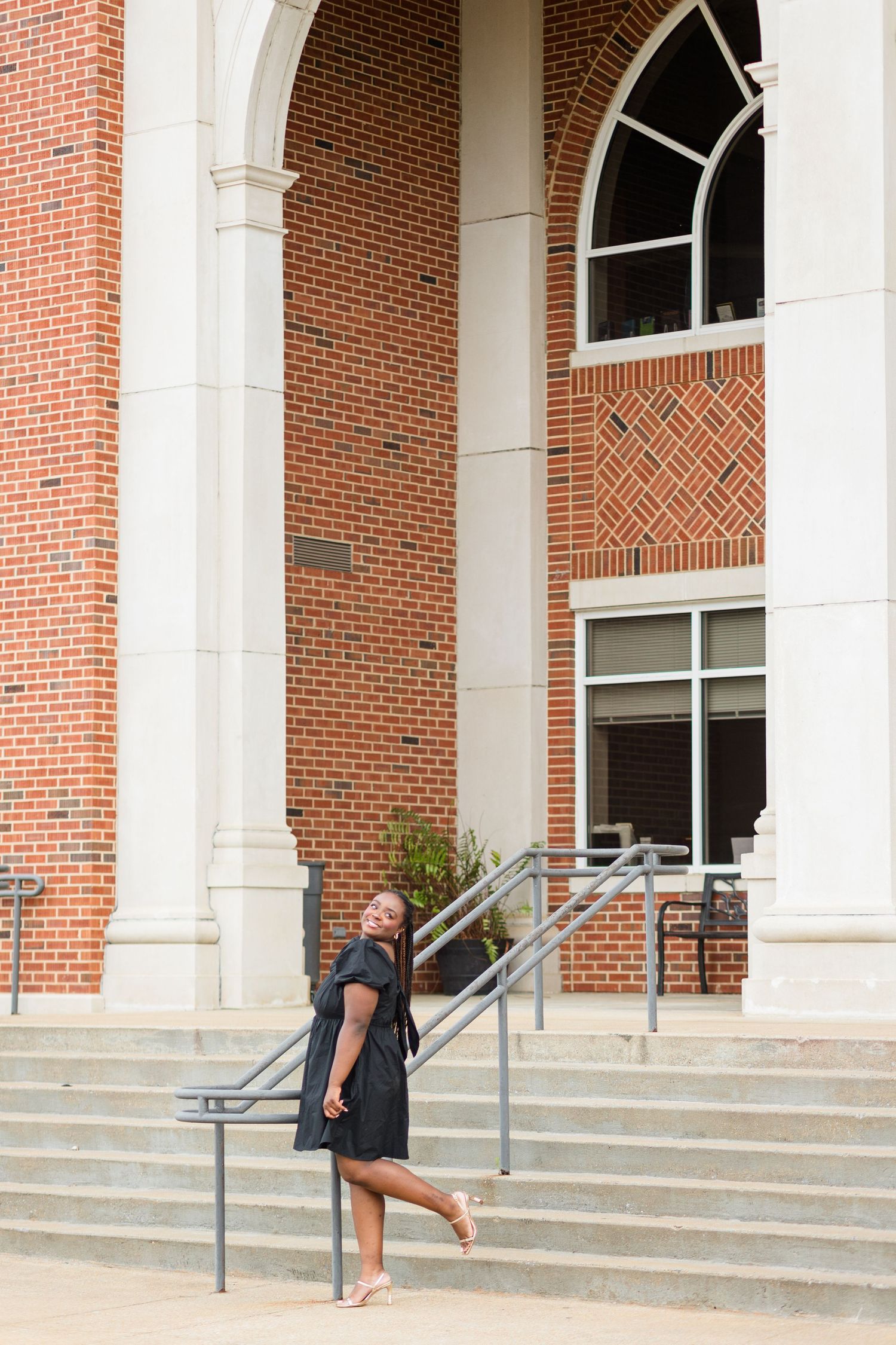 Young woman in black dress posing on steps of a red brick building with white columns.