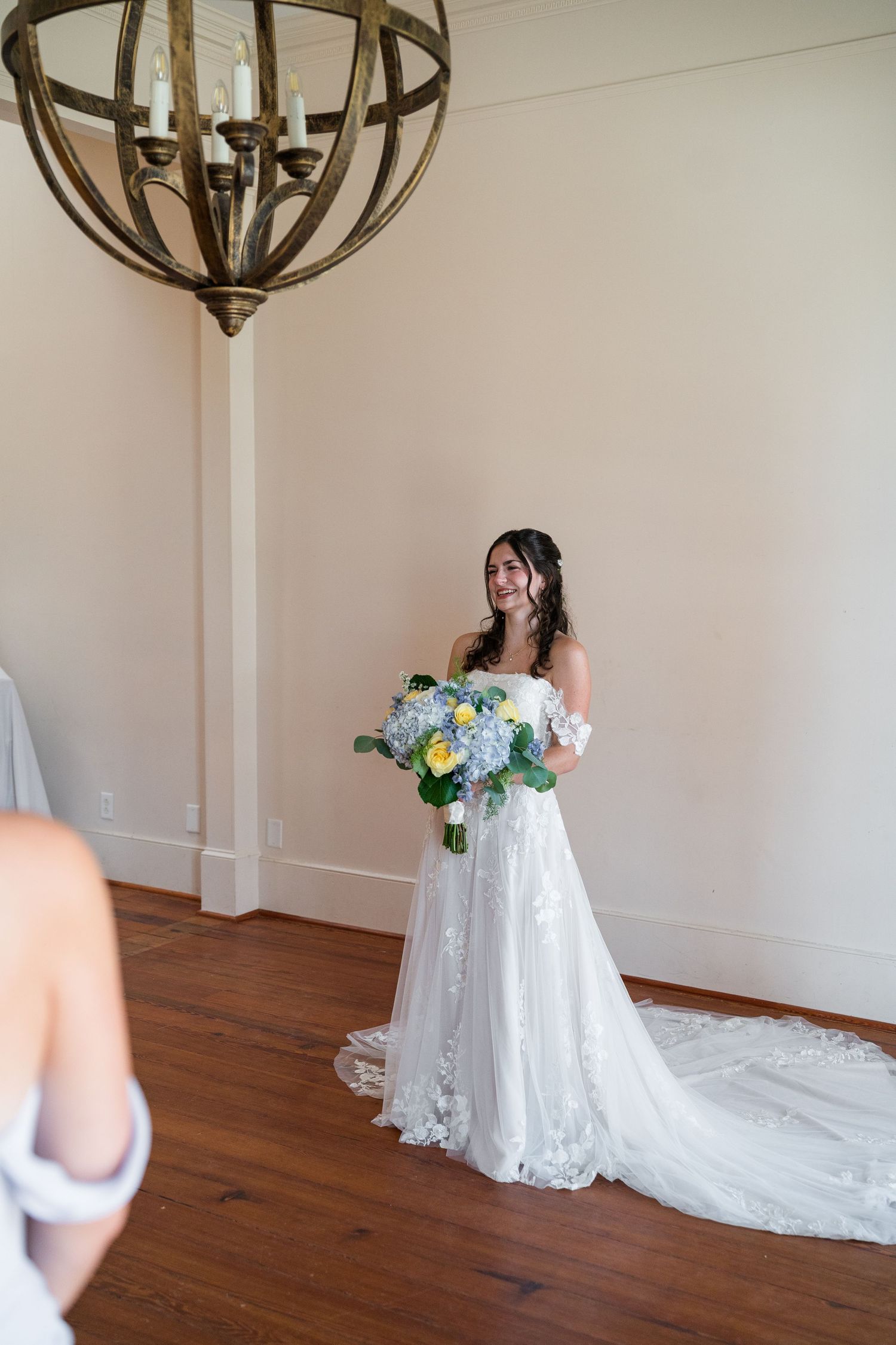 Bride in lace off-shoulder gown holds blue and yellow bouquet under chandelier in elegant venue.