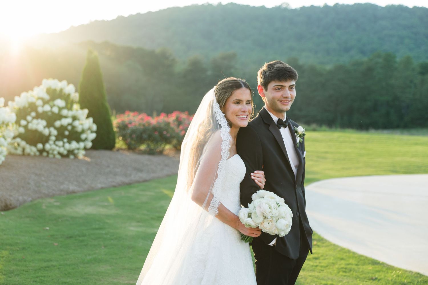 Bride and groom pose together on a golf course at sunset, she in a white gown with veil, he in a black tuxedo.