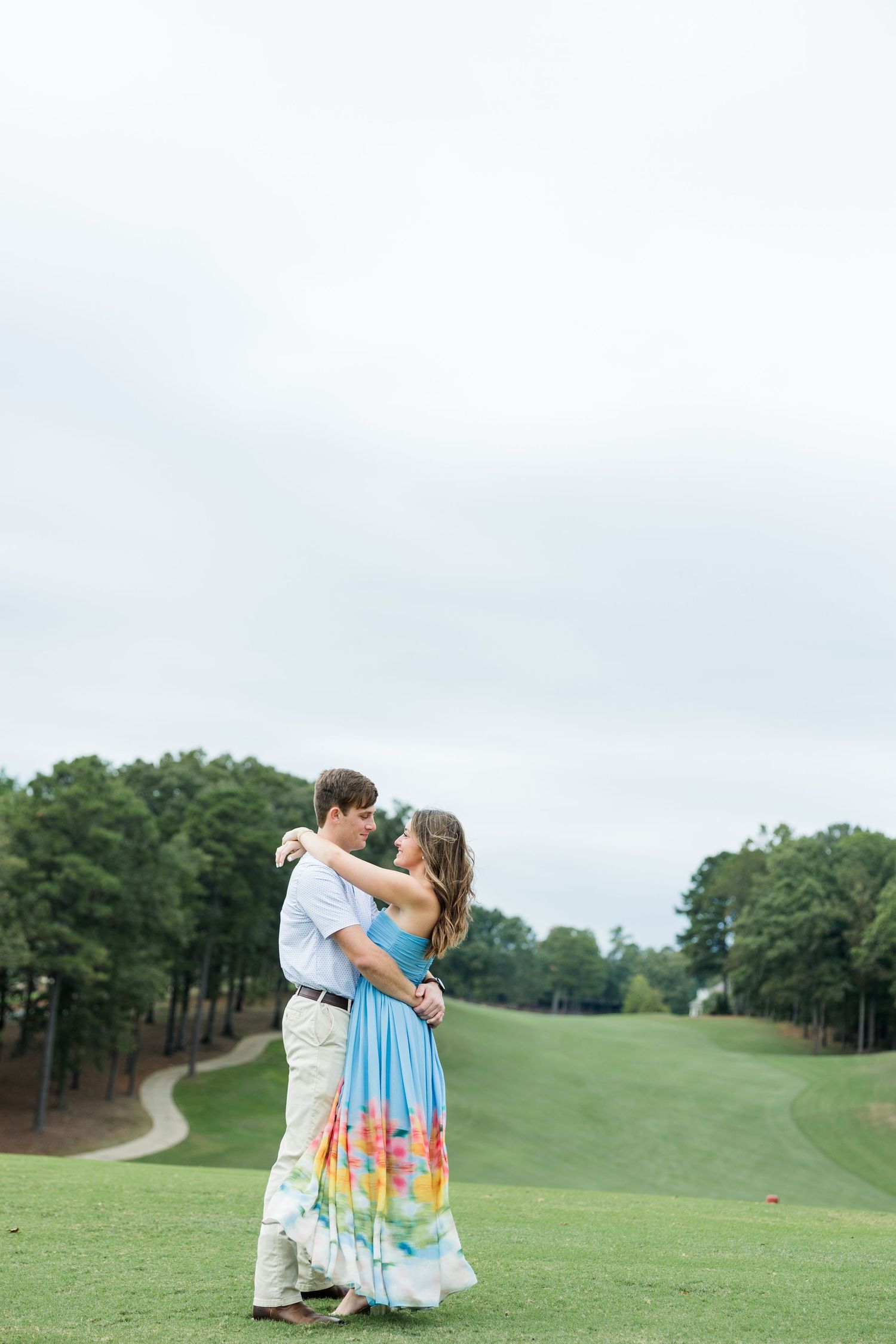 Couple embraces on a green golf course fairway, the woman in a colorful floral maxi dress, with tall pine trees under a cloudy sky.