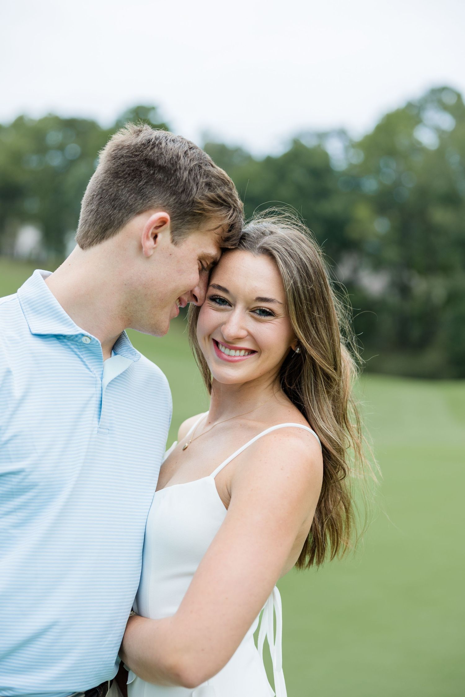A happy young couple shares an affectionate moment on a green lawn, the man touching his forehead to the smiling woman's head.