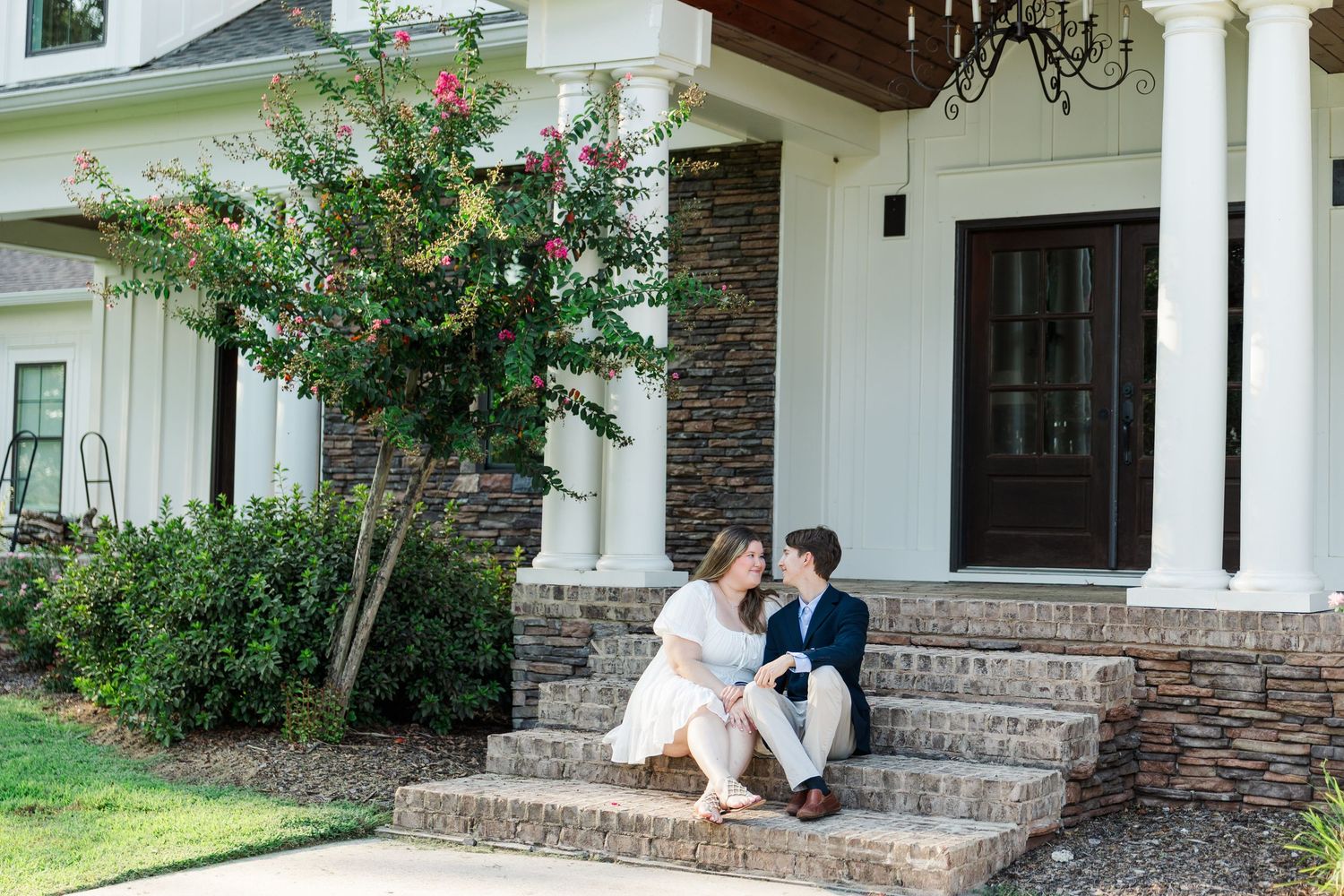 Couple sitting on front porch steps of elegant white home with stone accents and blooming pink crepe myrtle tree.