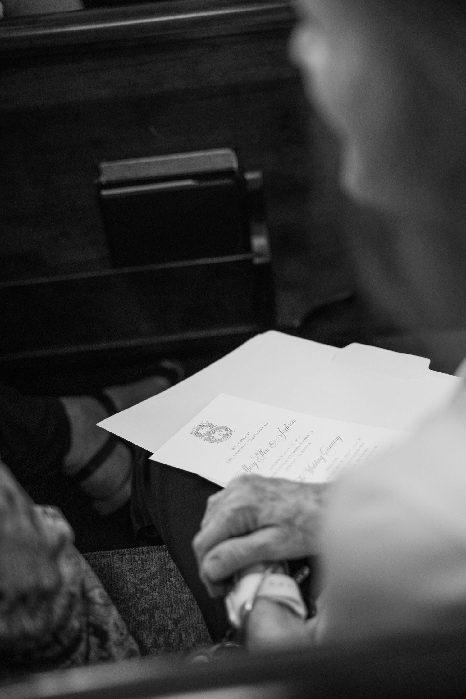 Black and white photo of wedding guest reading ceremony program on wooden church pew.