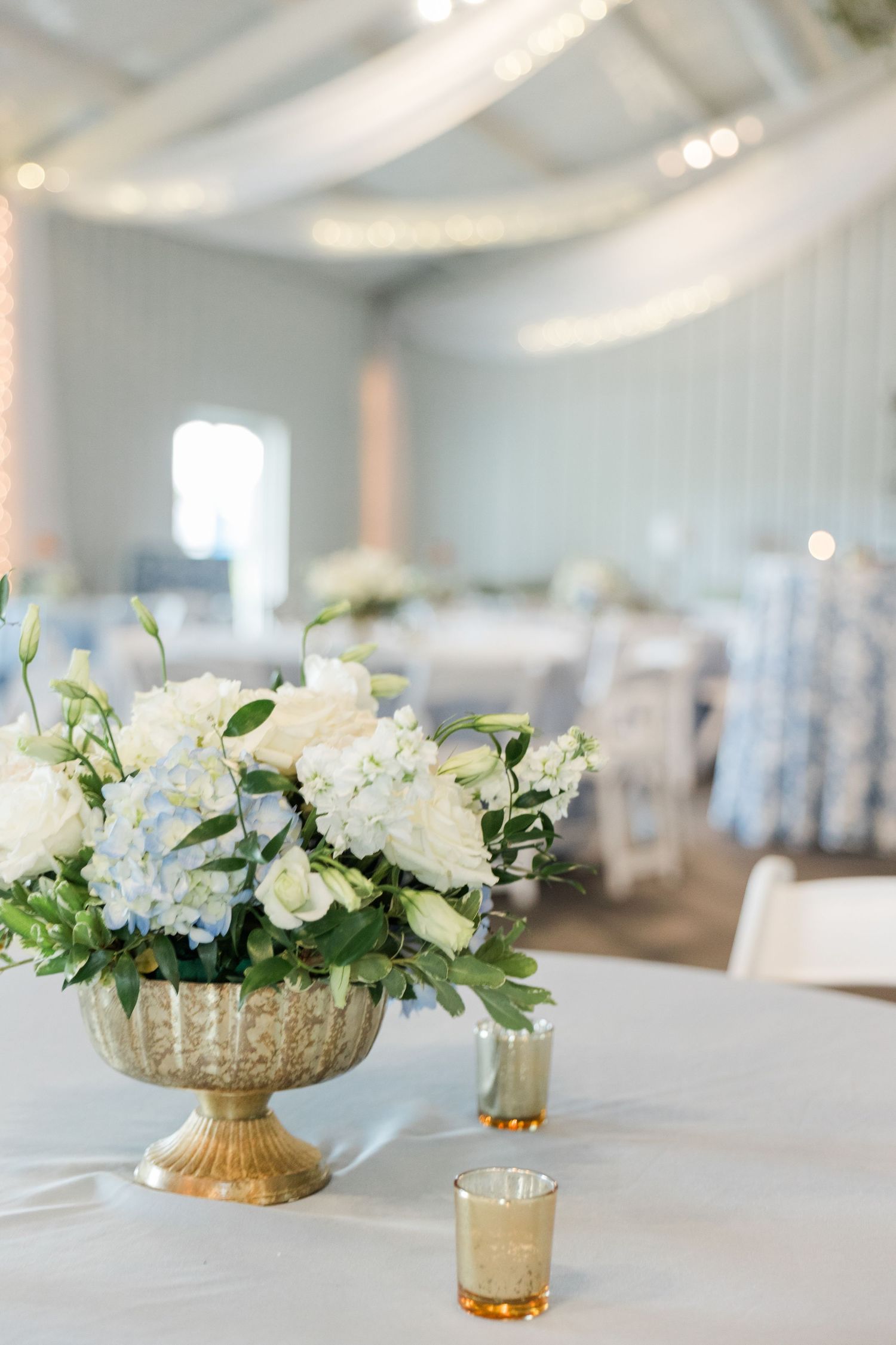 Gold urn centerpiece filled with white roses and blue hydrangeas on white tablecloth at wedding reception.