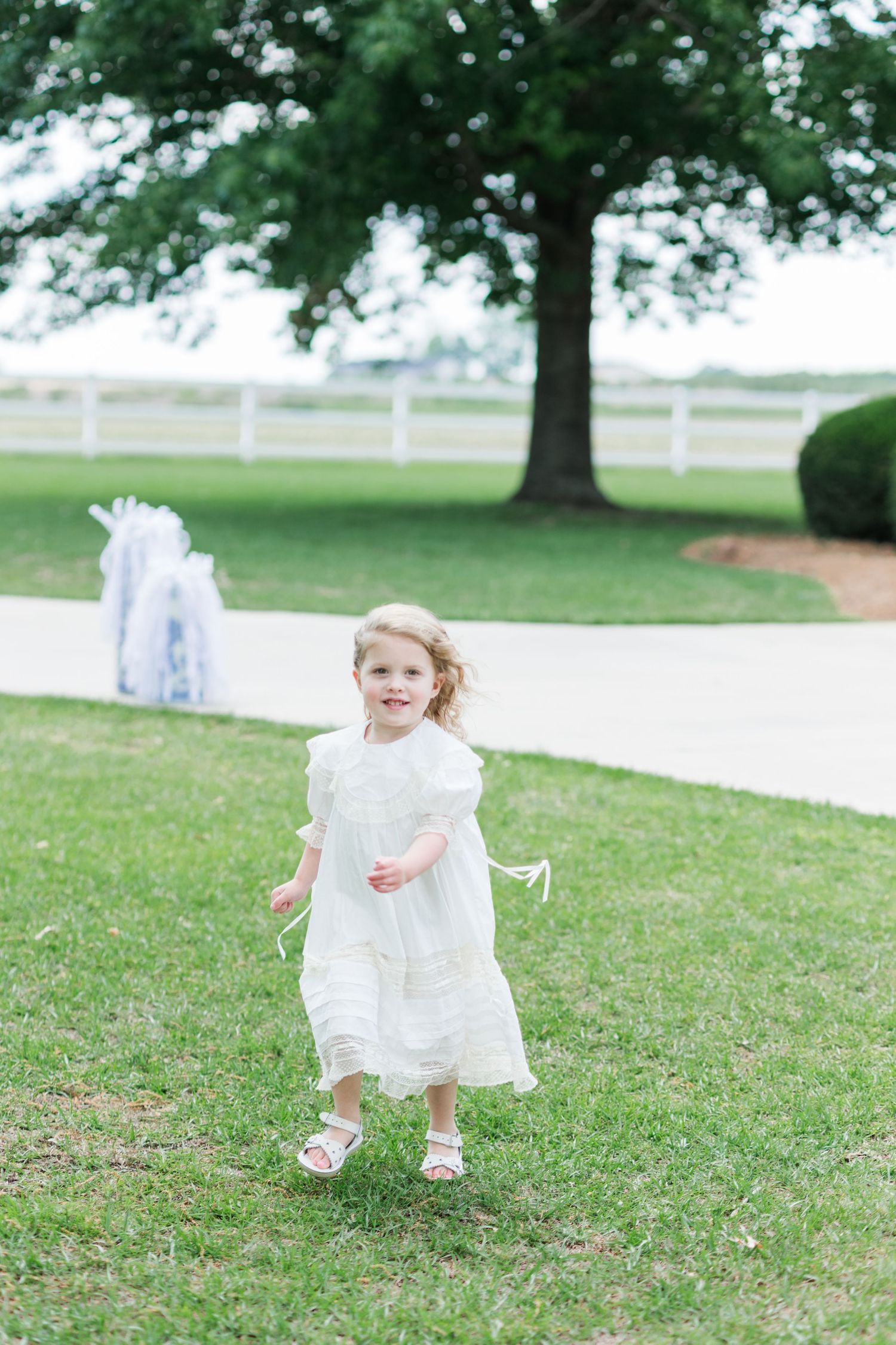 A young flower girl in a white lace dress runs across green grass at an outdoor wedding ceremony.