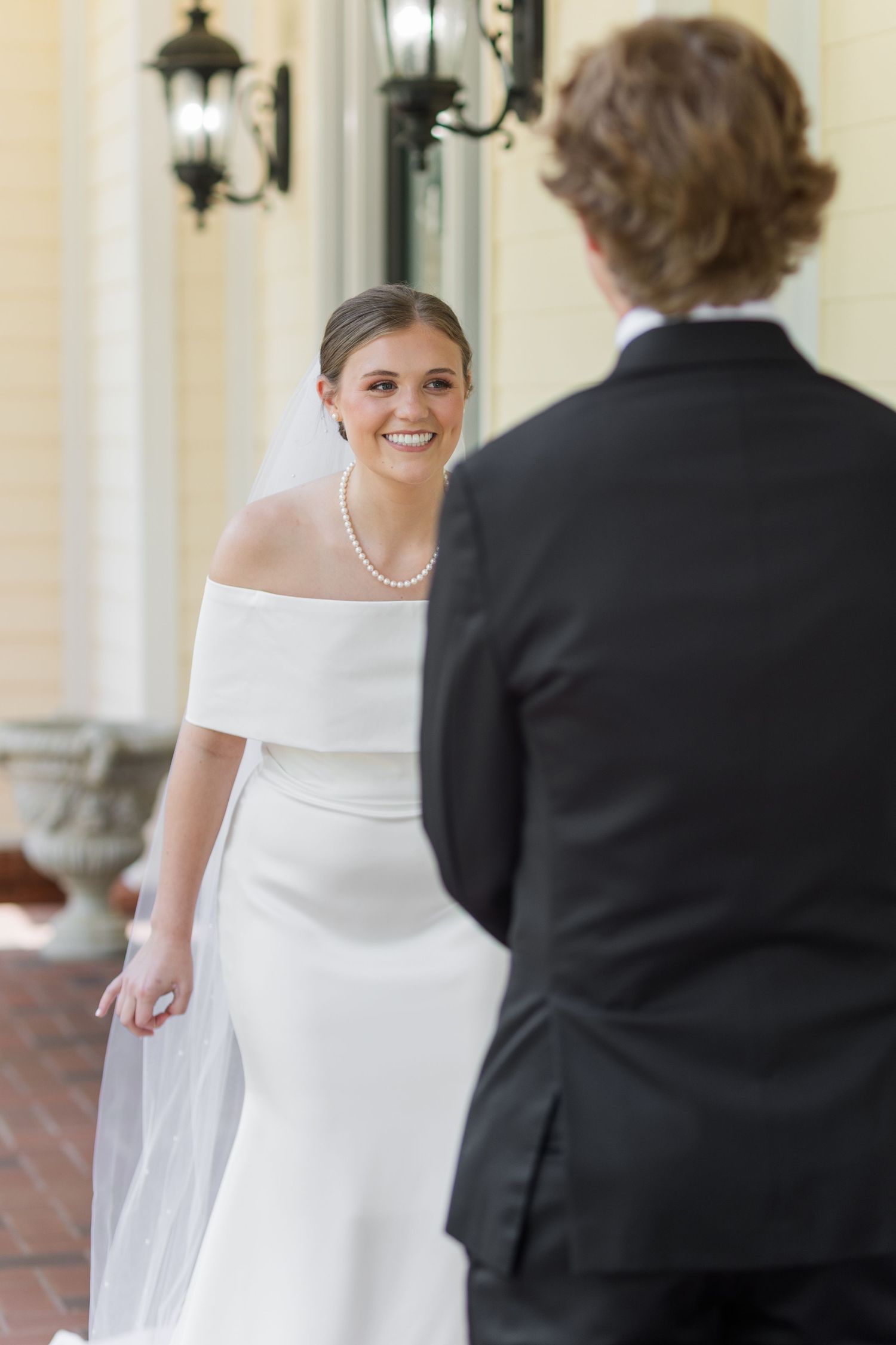 Smiling bride in off-shoulder white gown and pearl necklace during first look with groom.