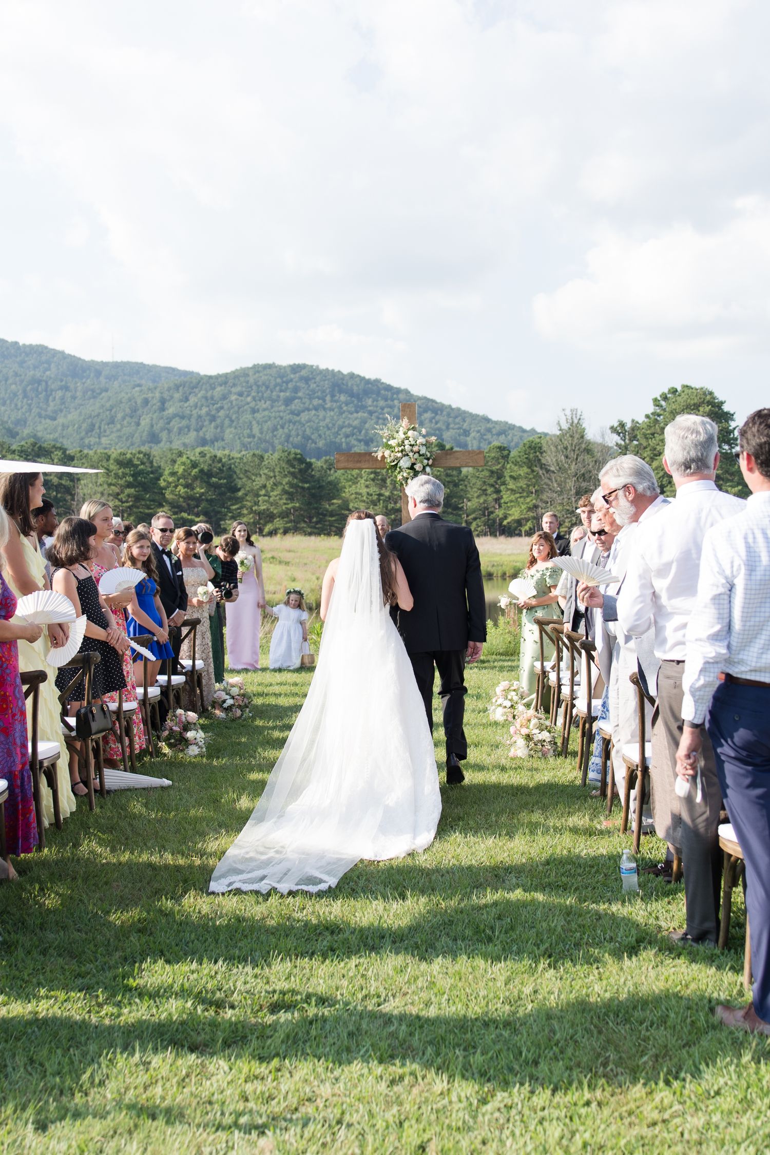 Bride walking down outdoor aisle with father at scenic mountain wedding ceremony with guests standing on green lawn.
