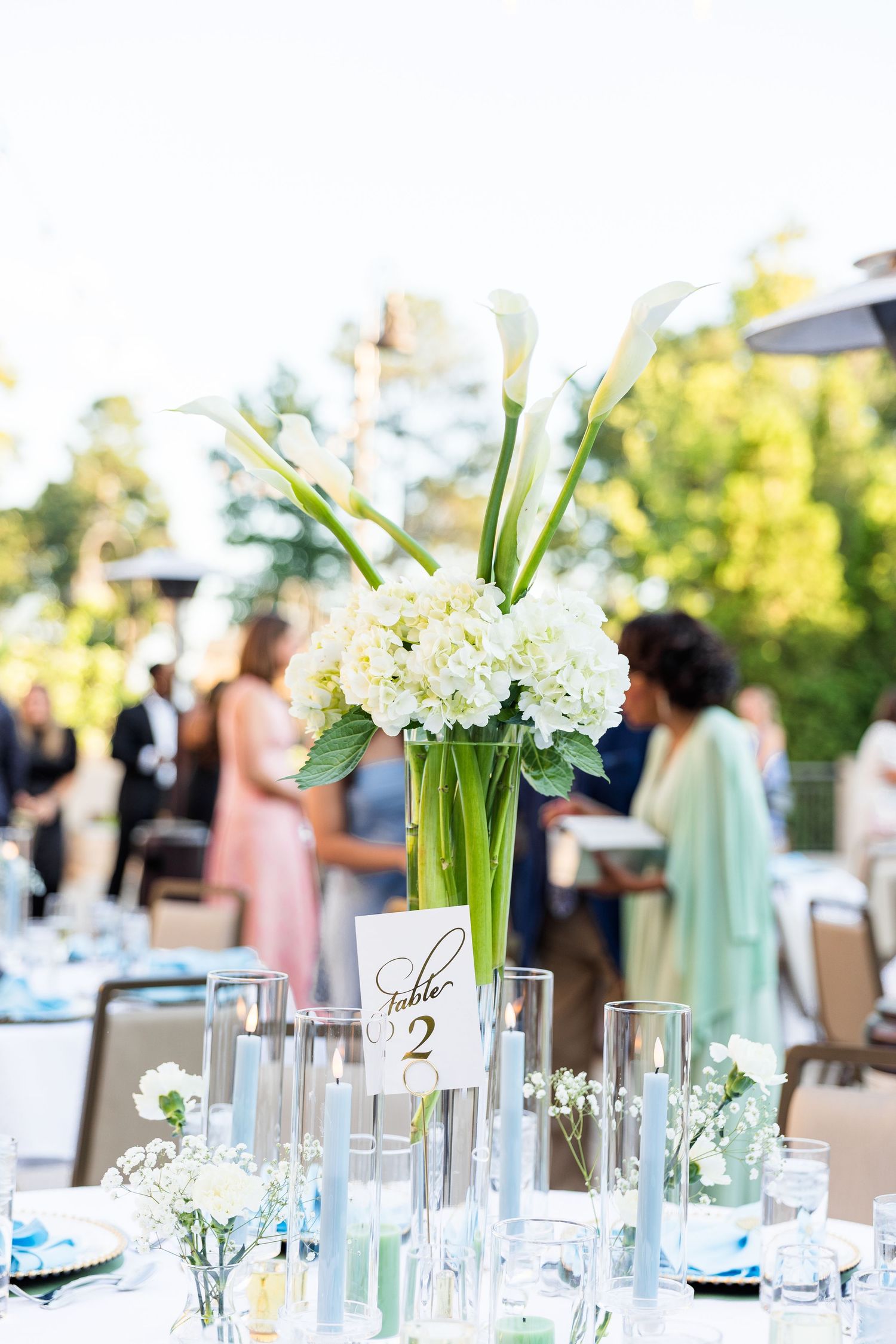 Elegant wedding reception table with white hydrangea centerpiece, calla lilies, candles, and Table 2 sign outdoors.
