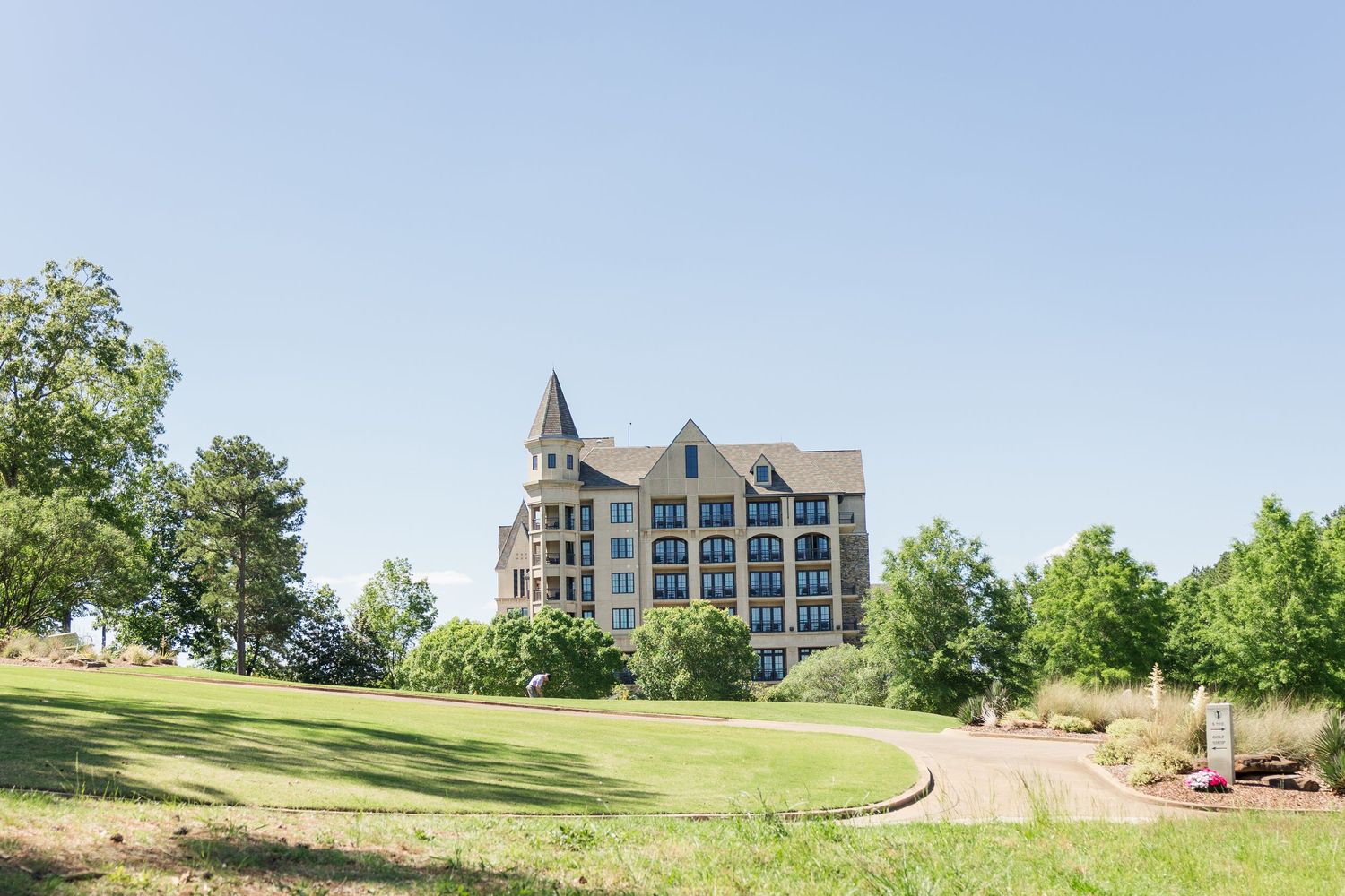 Large historic stone building with a turret sits atop a green hill surrounded by trees under a clear blue sky.