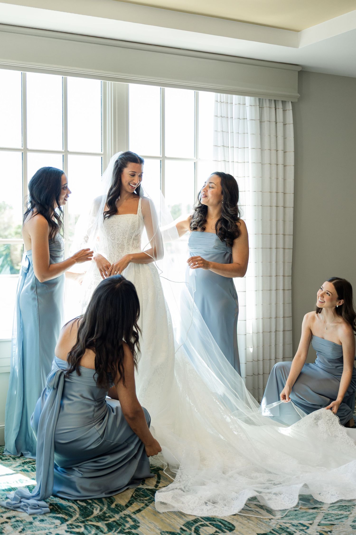 Bride in white gown with bridesmaids in dusty blue dresses helping her prepare near a bright window.