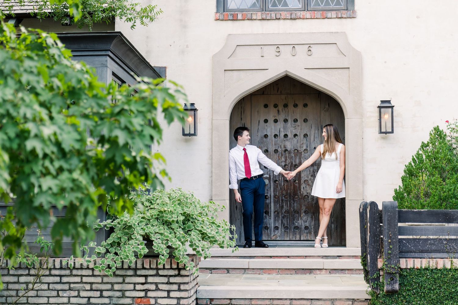 A couple holds hands on steps before a grand historic door, man in red tie and woman in white dress.