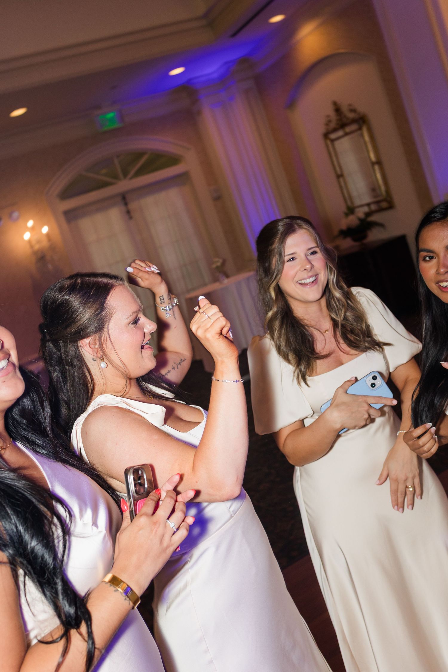 Women in white dresses dancing and smiling at an elegant wedding reception venue with purple uplighting.