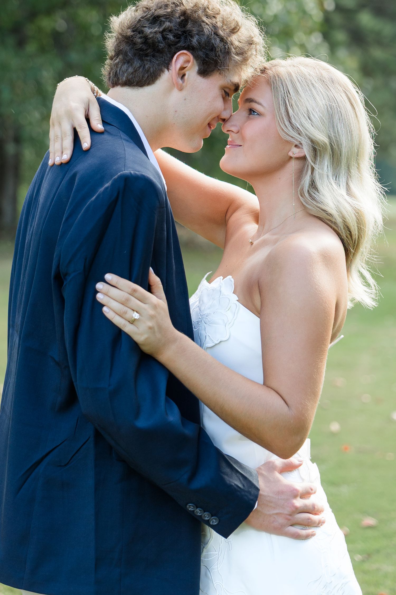 A bride and groom share a romantic moment outdoors, dressed in a white gown and navy suit, smiling closely together.