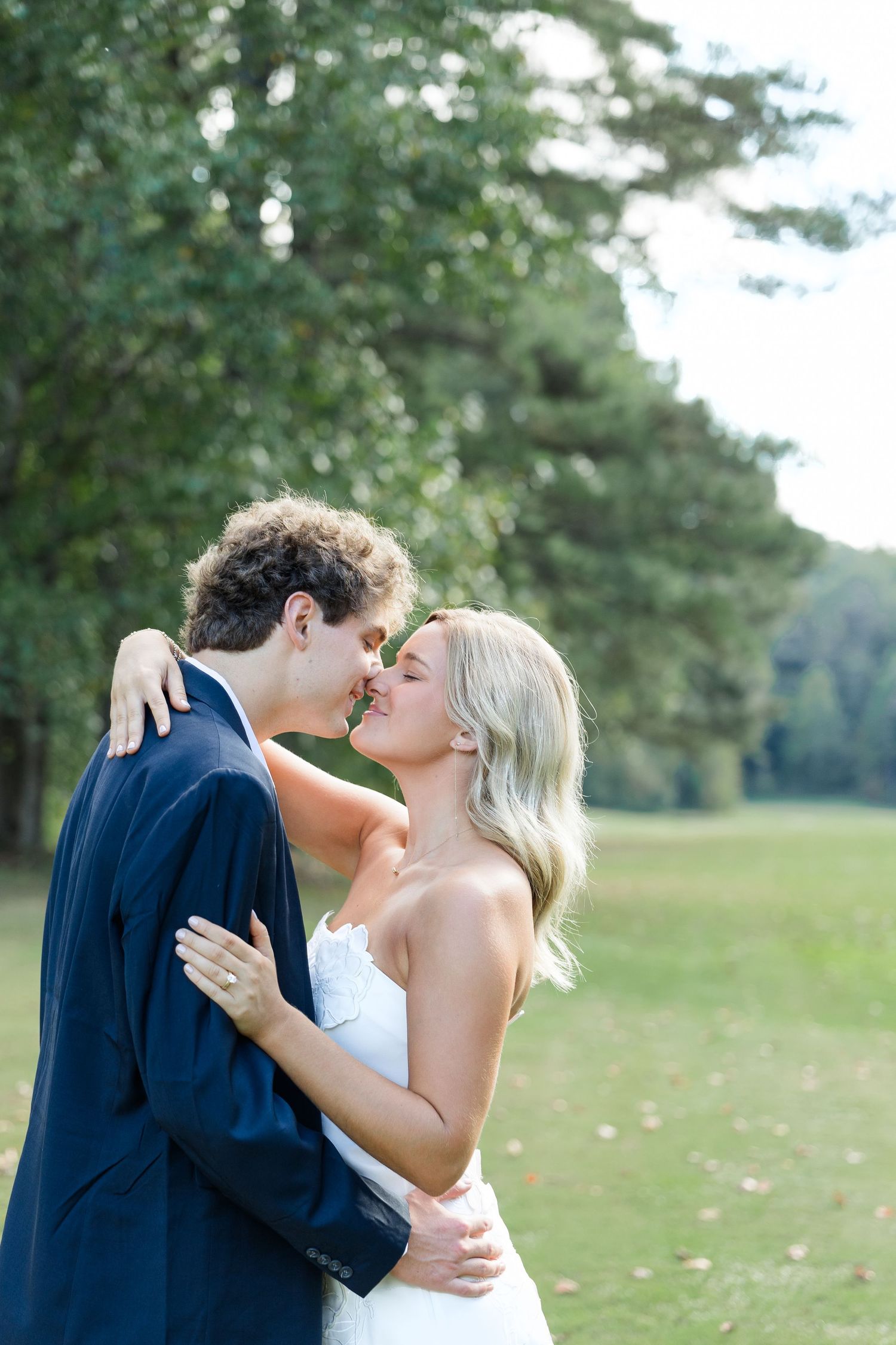 Bride and groom sharing a romantic almost-kiss outdoors on a green lawn with lush trees in the background.