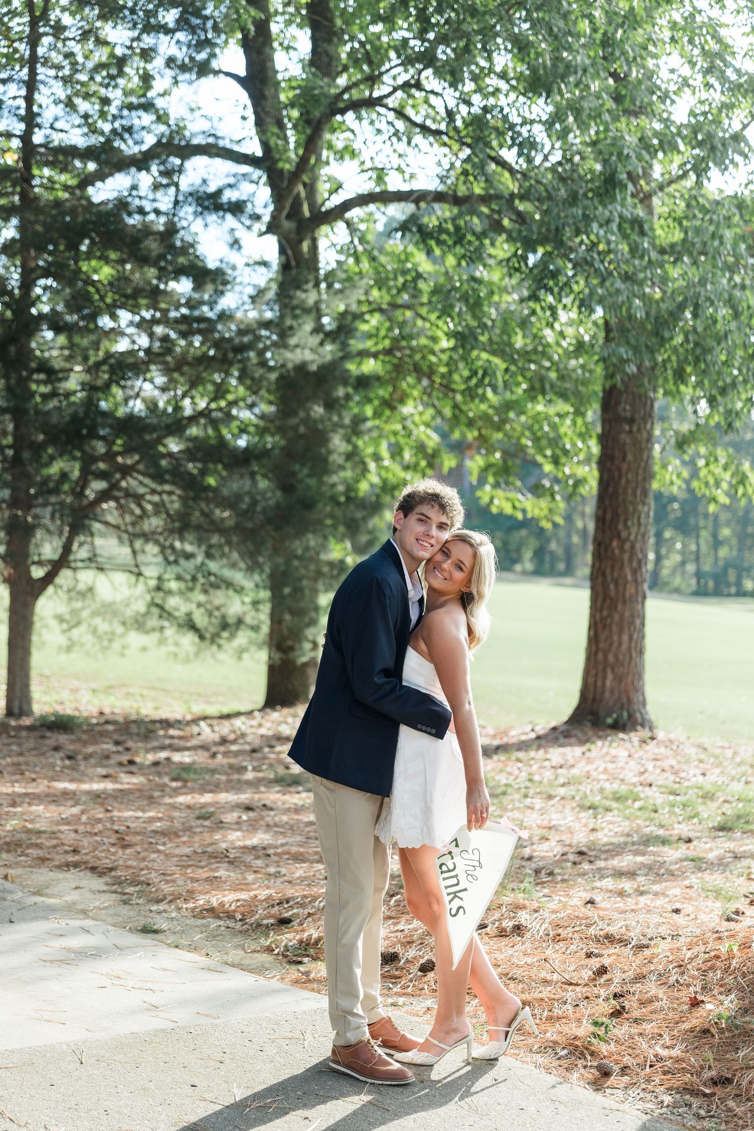 Couple embracing on a tree-lined path, woman in white dress holding a decorative sign outdoors.