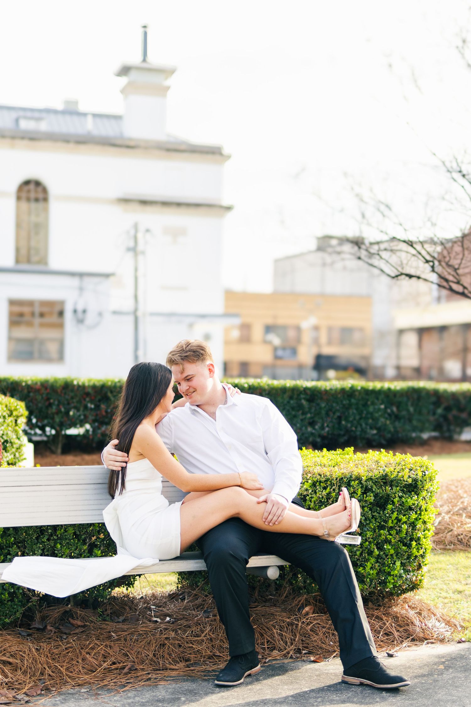 Couple sitting closely on a white bench in a sunny park, woman in white dress with legs on man's lap.