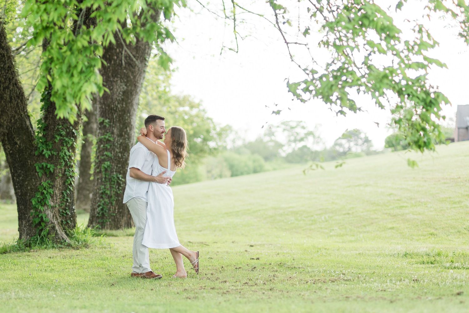 A couple shares a romantic kiss under a lush green tree in an open grassy field during an outdoor engagement session.