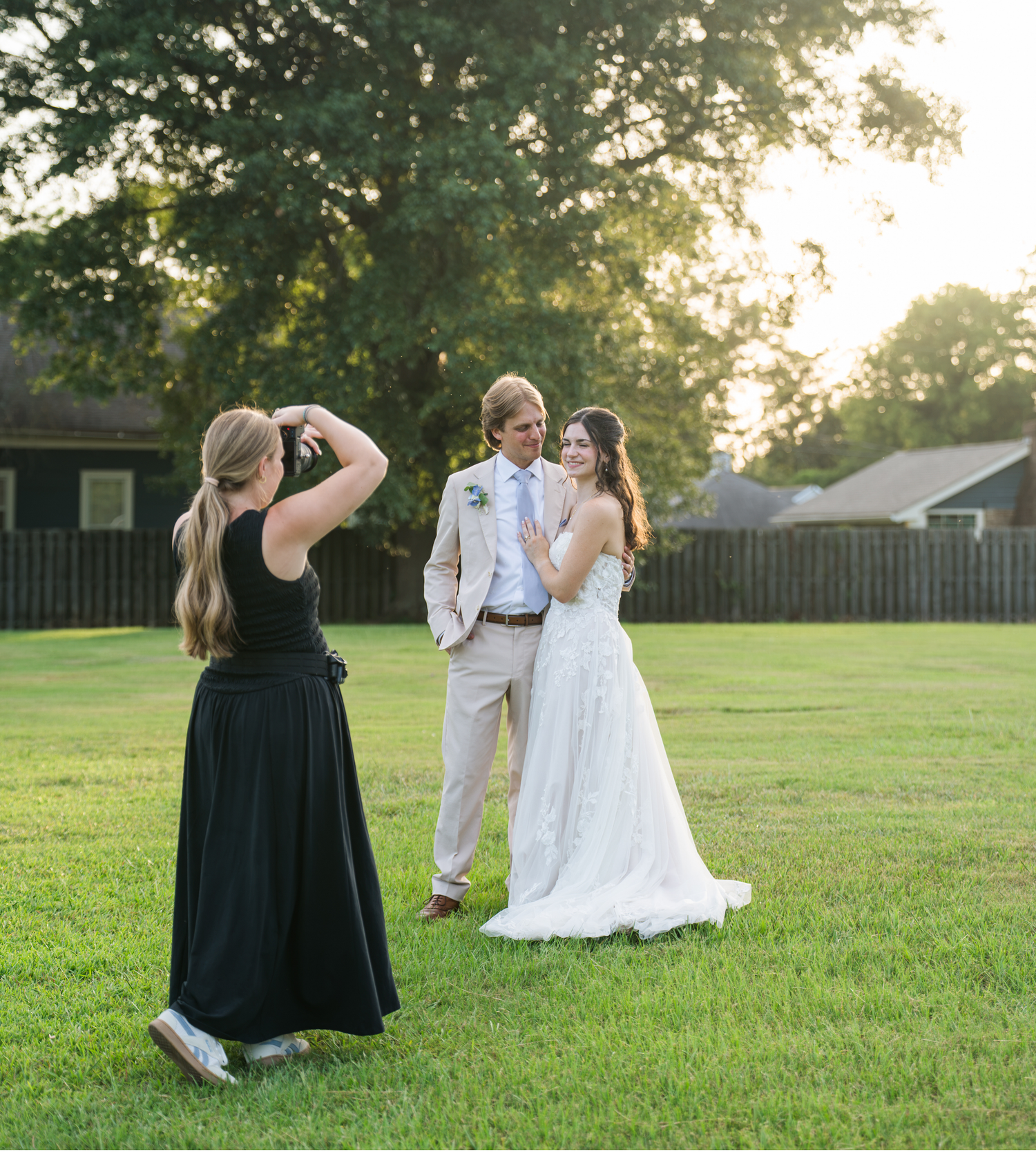 Photographer capturing bride and groom posing outdoors on green lawn at golden hour sunset wedding.