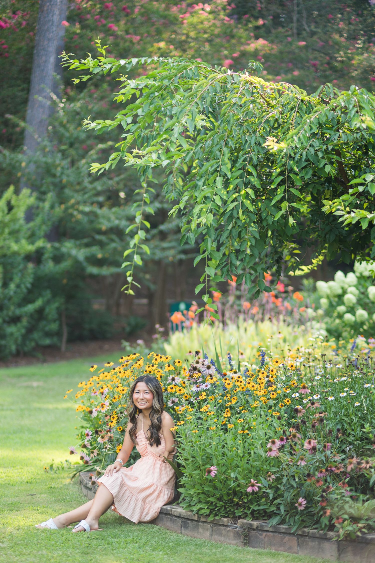 Young woman in pink dress sitting beside a colorful garden with yellow flowers and lush greenery.