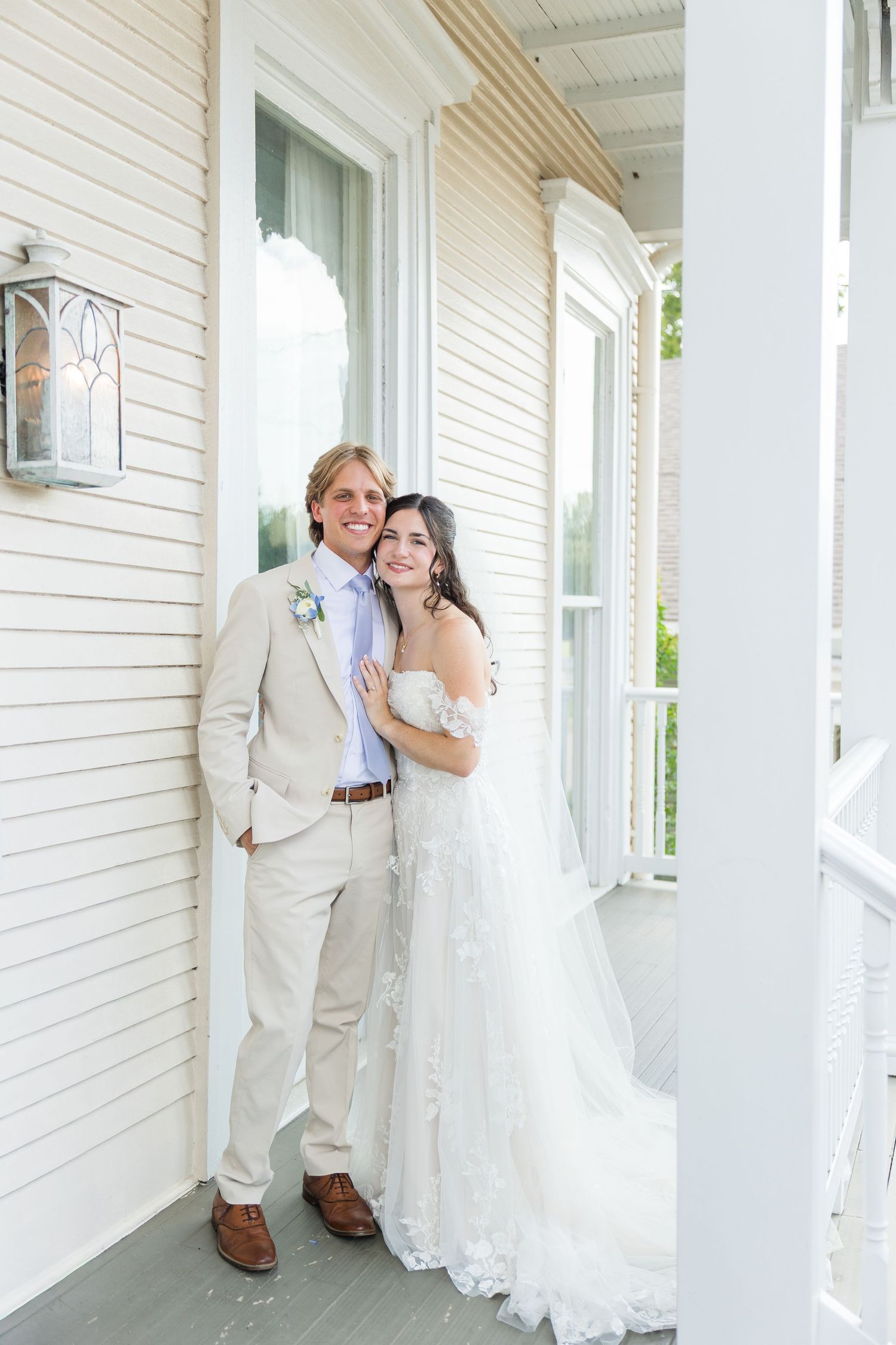 Happy bride and groom posing together on white porch, bride in lace gown, groom in tan suit.