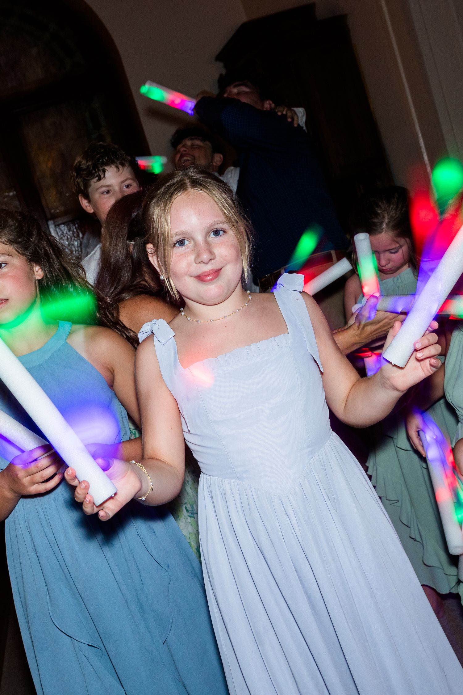 Young flower girl in light blue dress dancing with glowing foam sticks at a festive wedding reception.