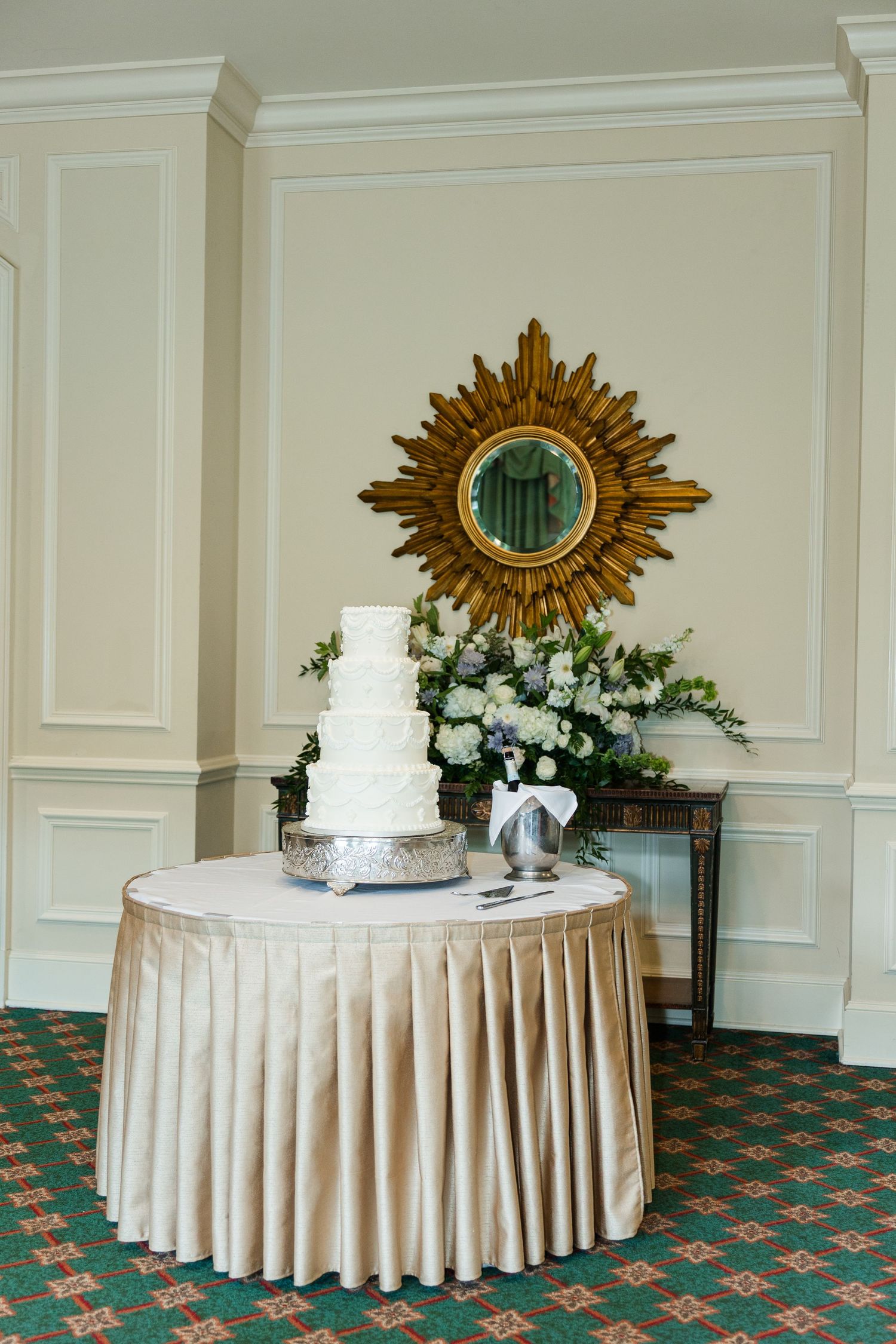 Elegant four-tier white wedding cake on a draped table with floral arrangement and gold sunburst mirror backdrop.