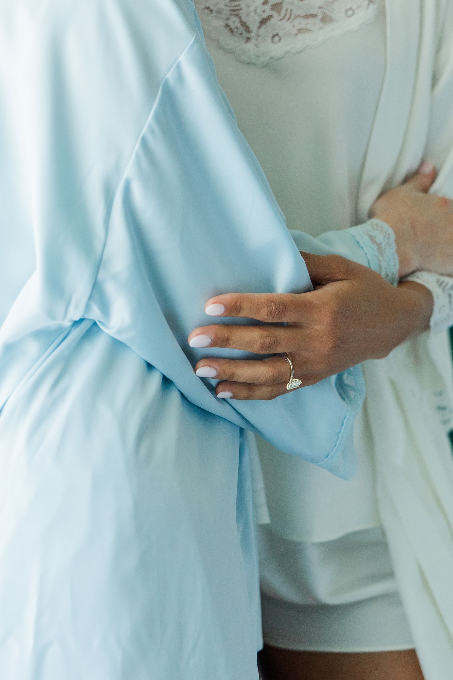 A bride in a light blue satin robe displays her diamond engagement ring while holding fabric close to her body.