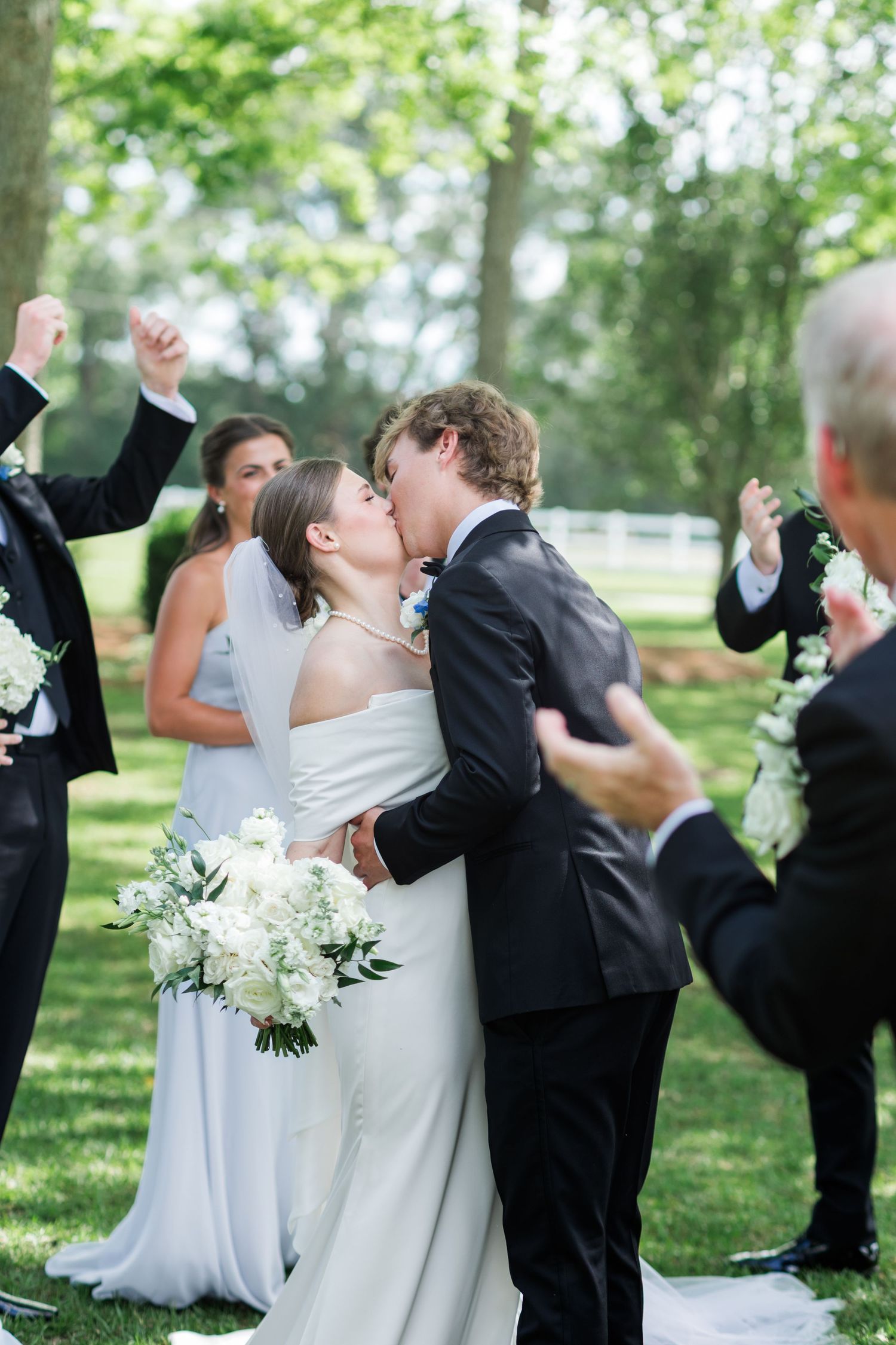 Bride and groom sharing first kiss at outdoor wedding ceremony surrounded by cheering guests.