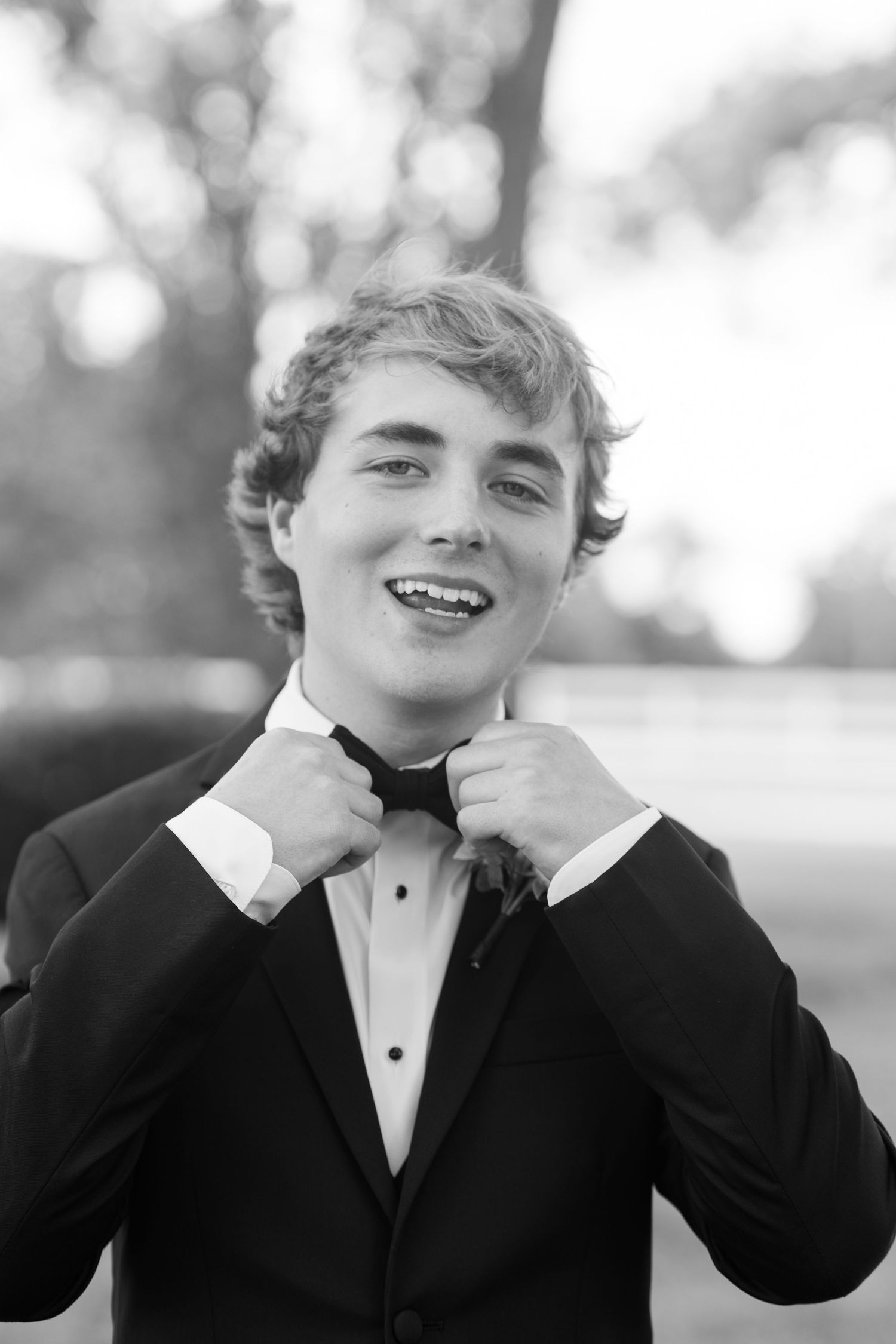 Young man in tuxedo adjusting bow tie, smiling in black and white portrait outdoors.