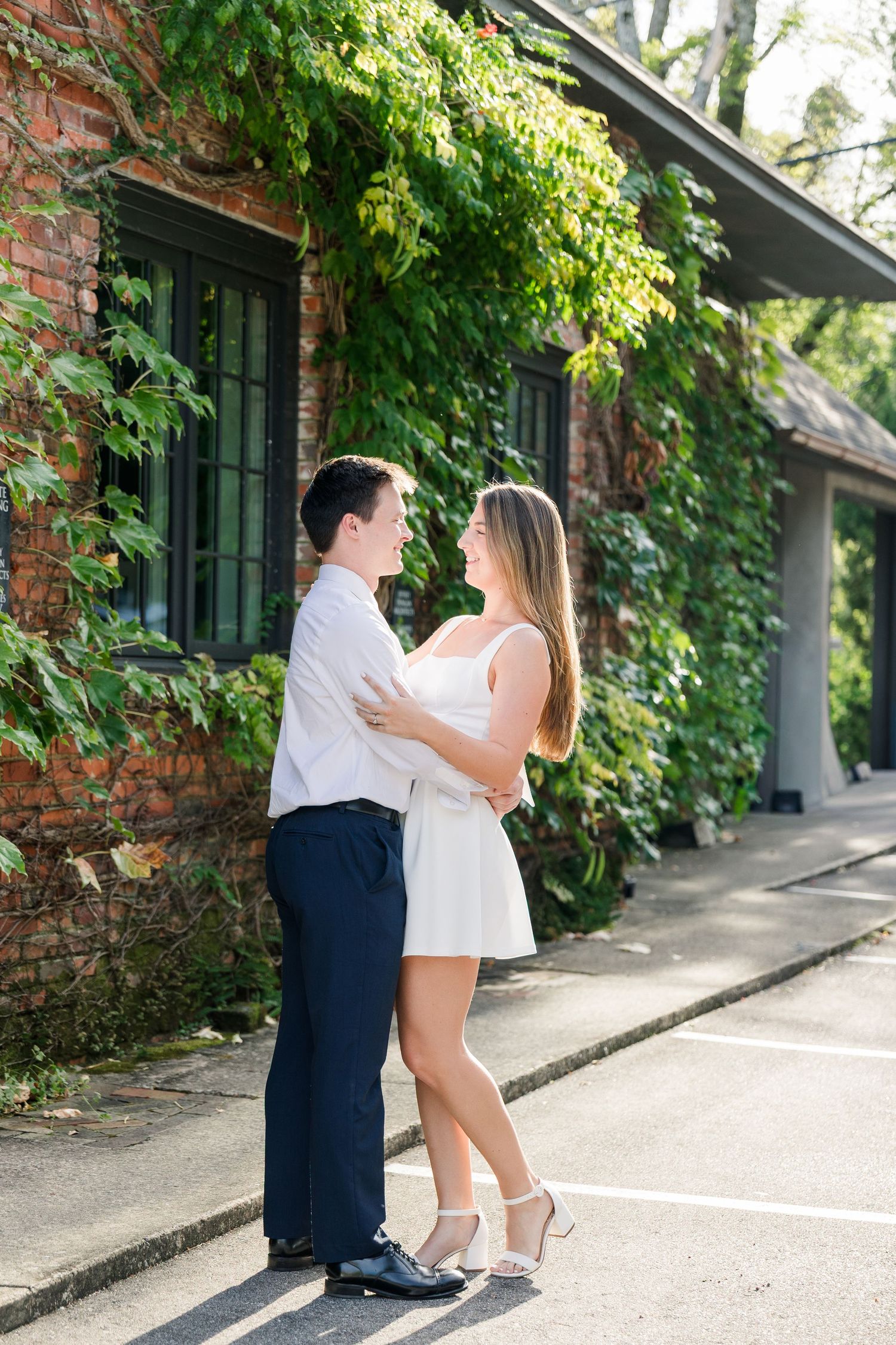 Couple embracing and smiling at each other outside an ivy-covered brick building on a sunny day.