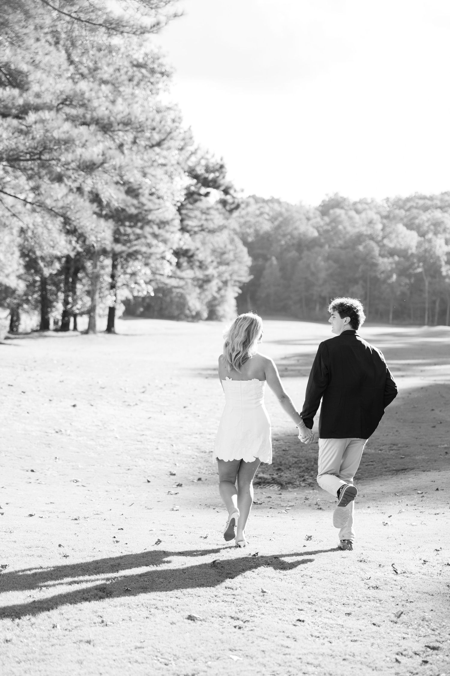 Black and white photo of a couple holding hands walking down a sunlit path surrounded by trees.