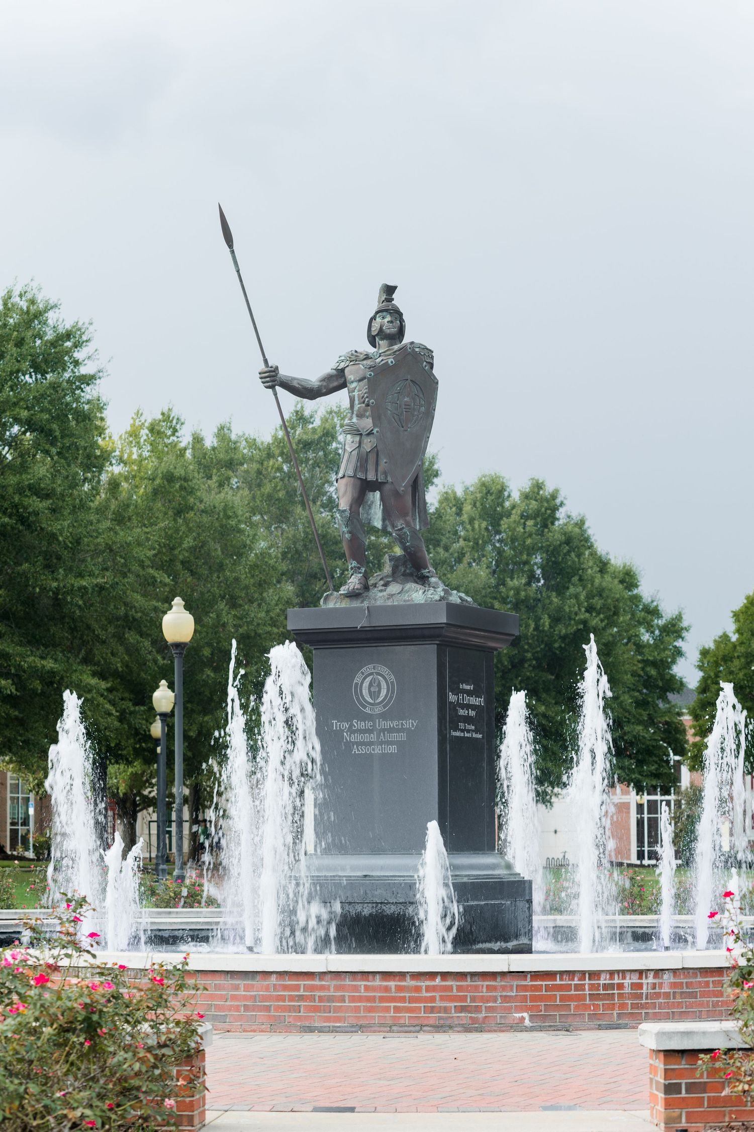 Bronze warrior statue with spear and shield stands on pedestal surrounded by fountains on a university campus.
