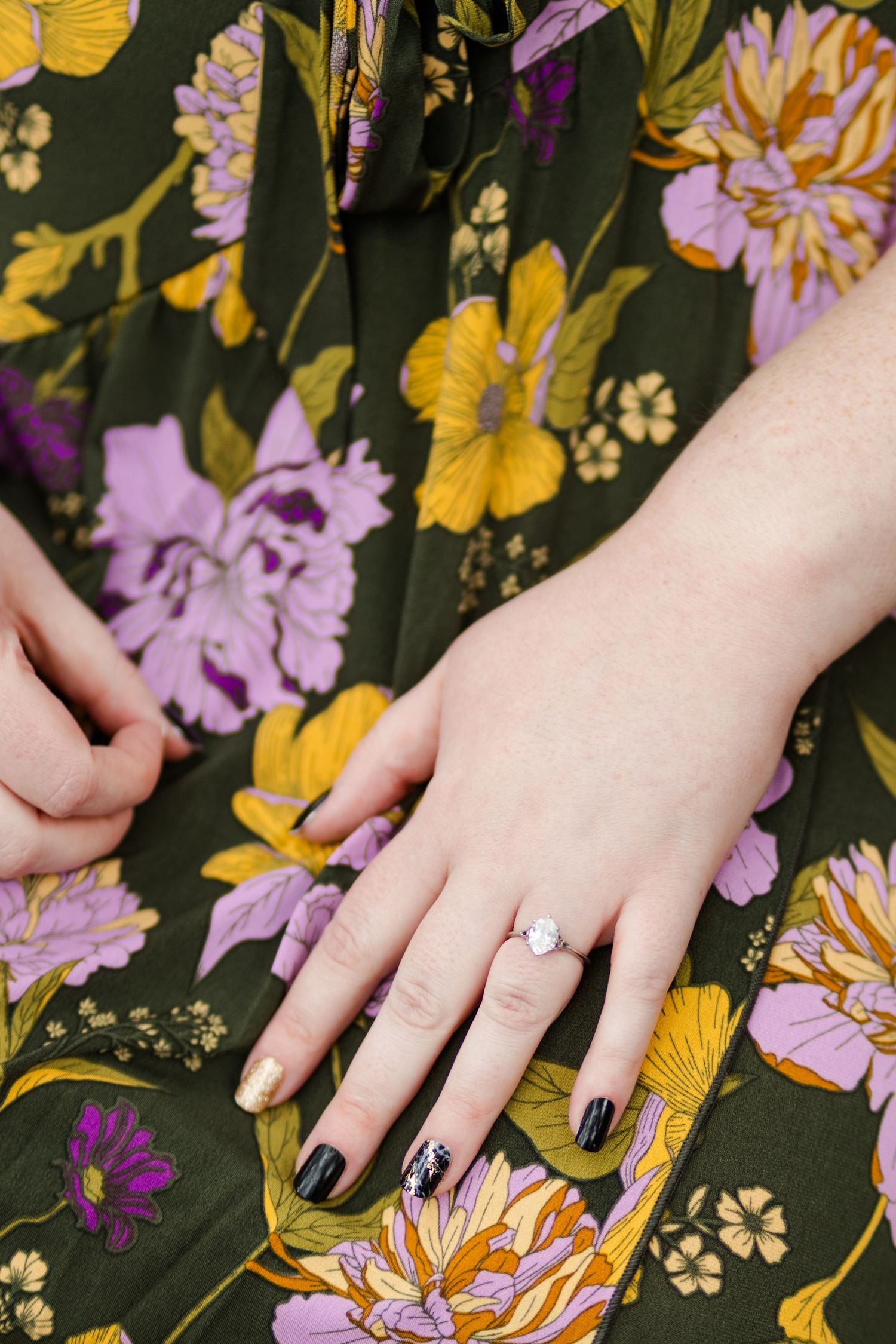 Close-up of hands wearing a white opal ring and black nail polish on a dark floral dress.