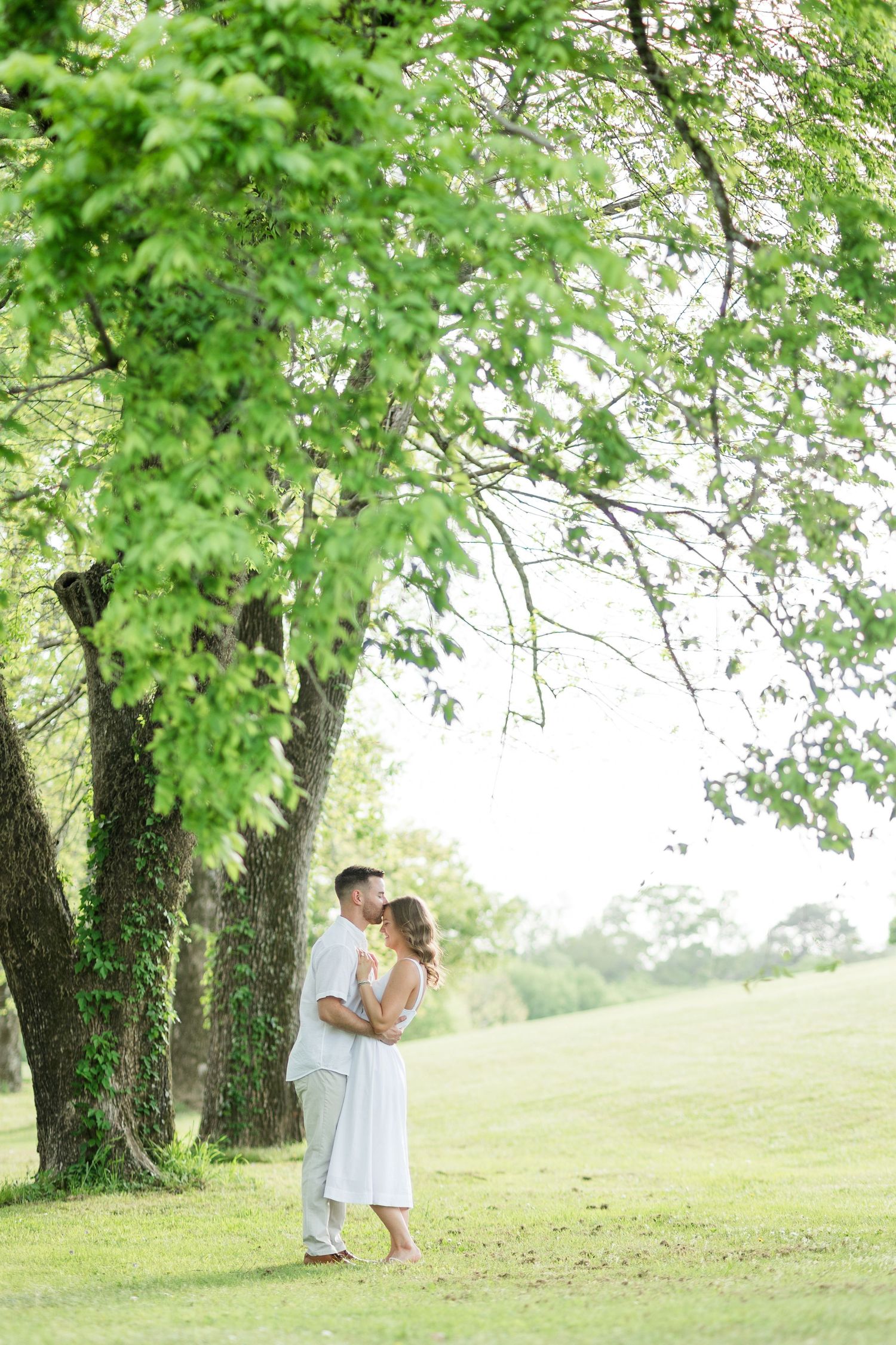Couple embracing and kissing under lush green trees in a sunlit park, wearing white outfits on a beautiful summer day.