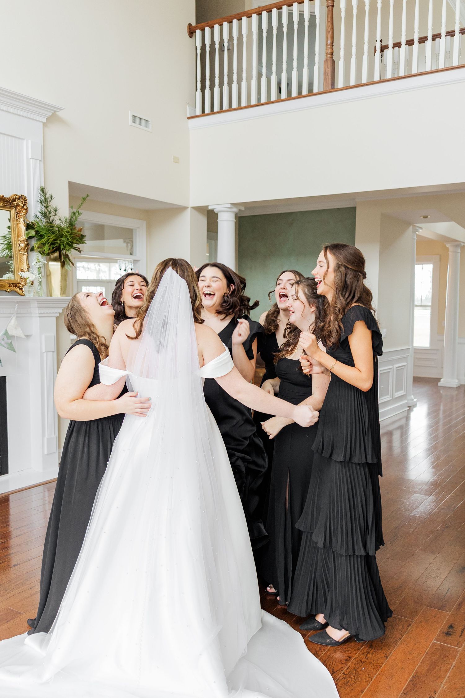 Bride in white gown and veil surrounded by bridesmaids in black dresses reacting with joy inside elegant home.