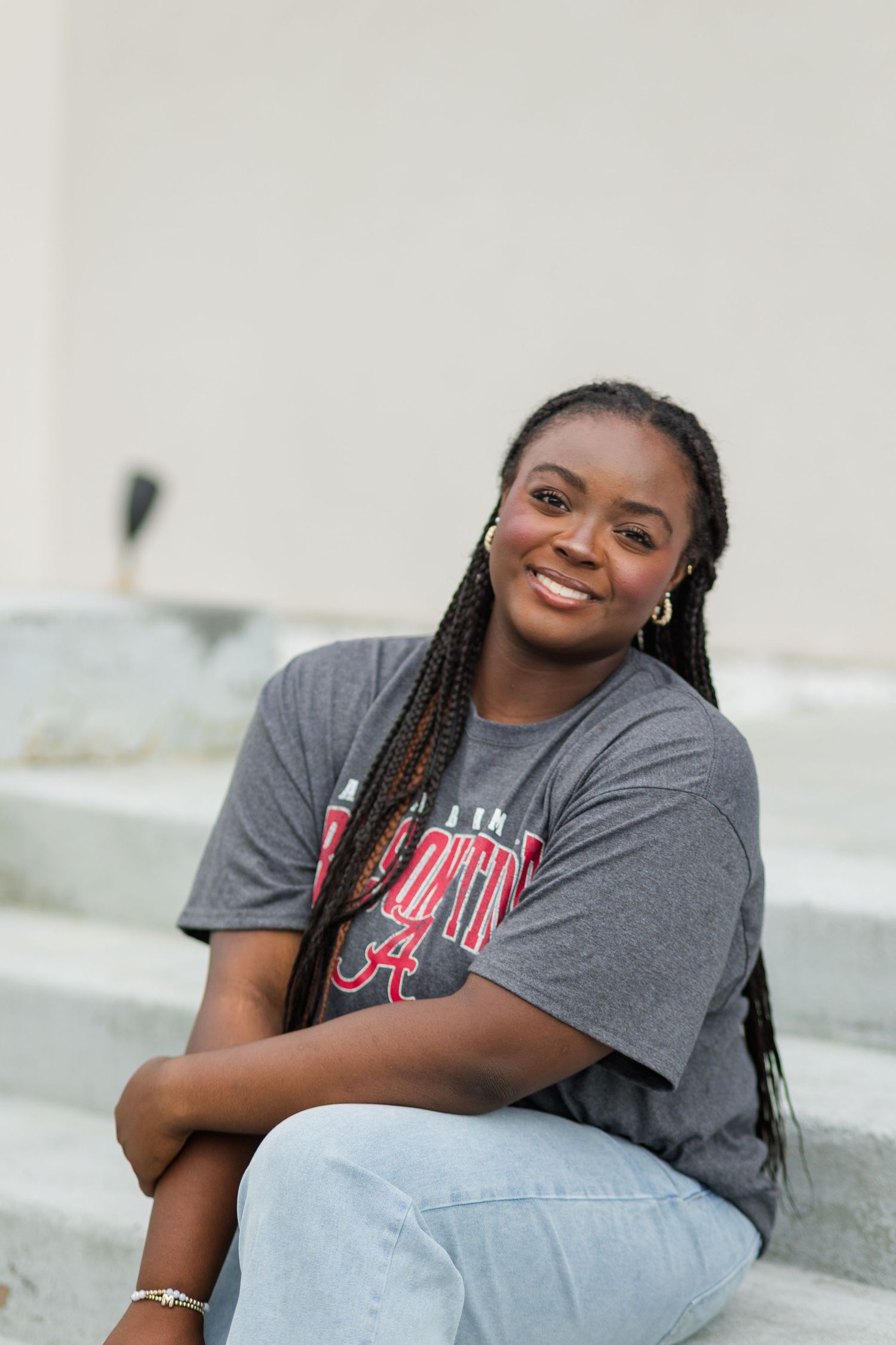 Young woman smiling in Alabama Crimson Tide shirt, sitting on steps with braided hair and jeans.