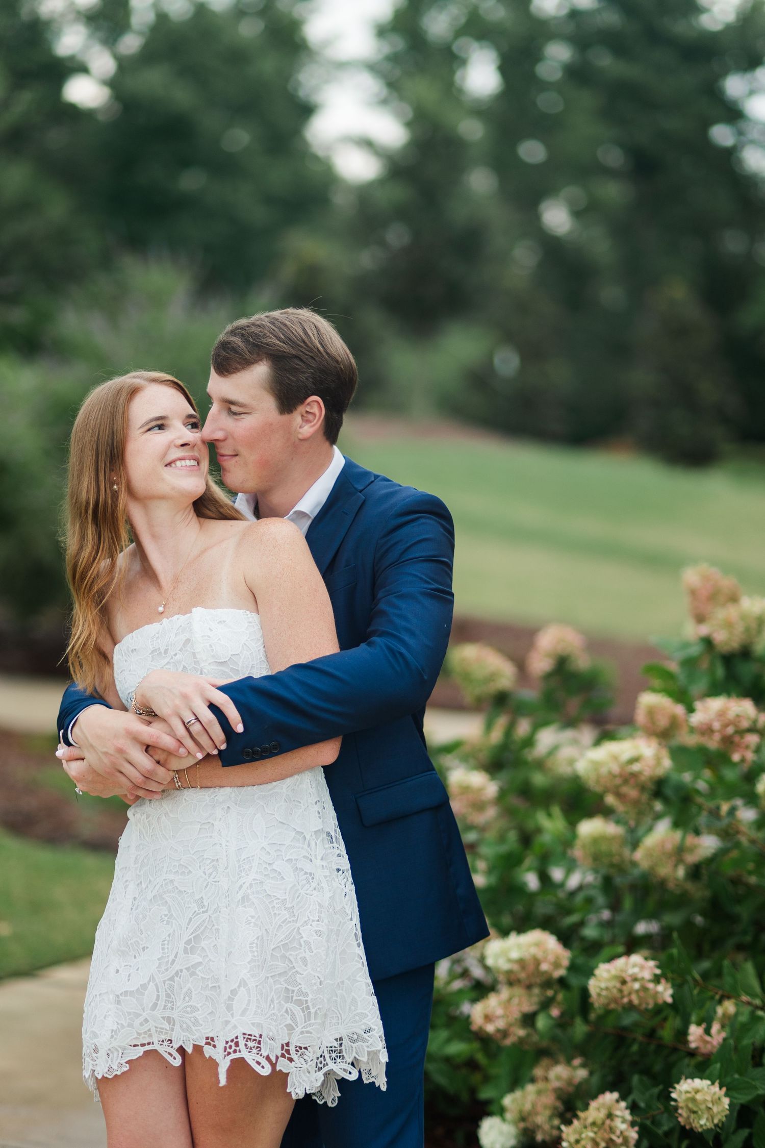 Couple embracing outdoors near blooming hydrangeas, woman in white lace dress and man in navy suit, smiling romantically.