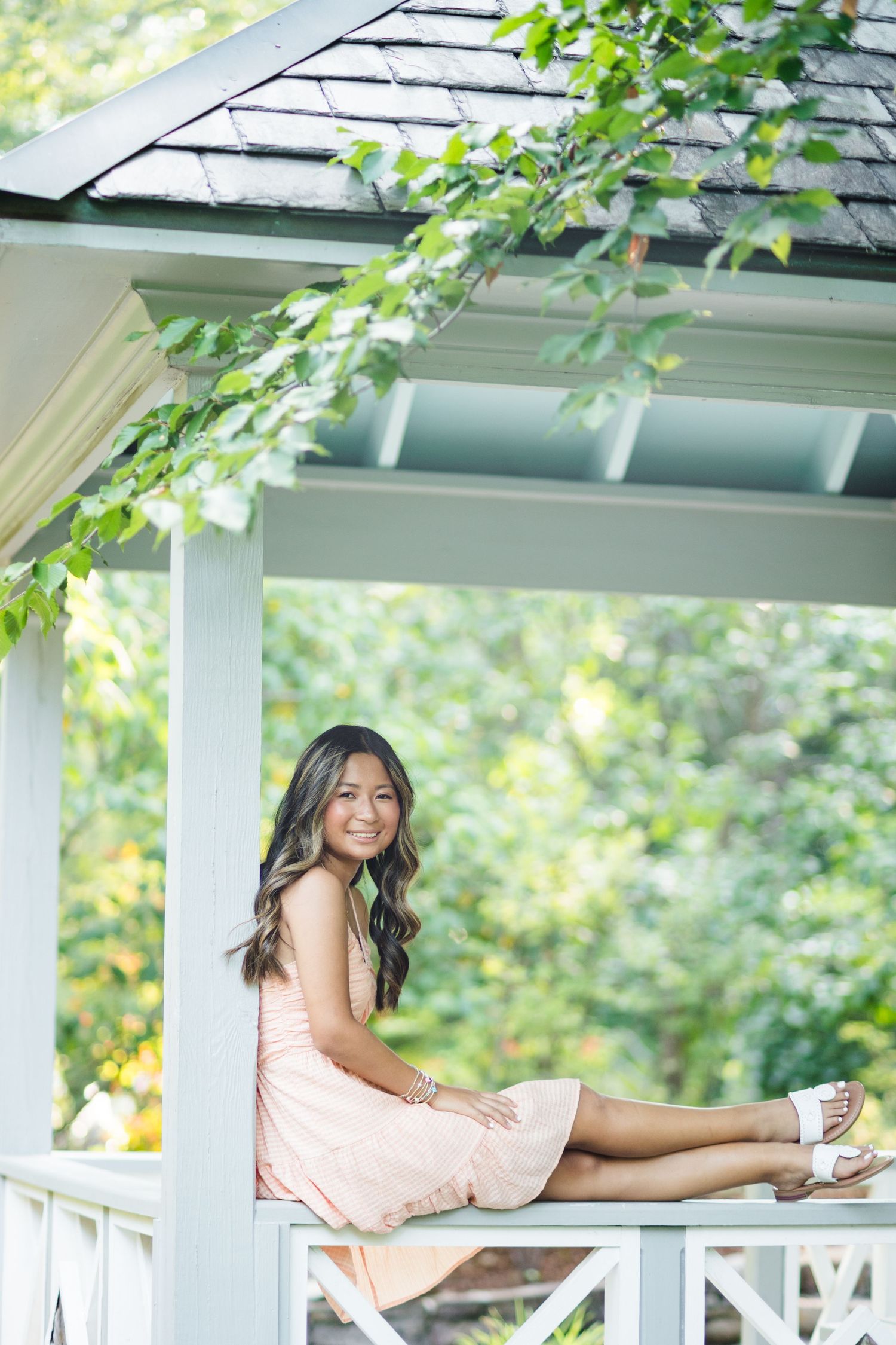 Smiling young woman in a peach dress sitting on a white gazebo railing surrounded by greenery.