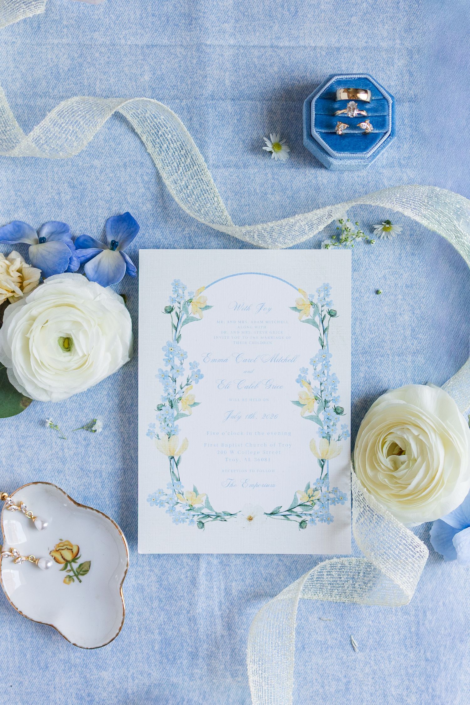 Blue floral wedding invitation suite with rings, white ranunculus, hydrangeas, ribbon, and vintage dish on blue linen.