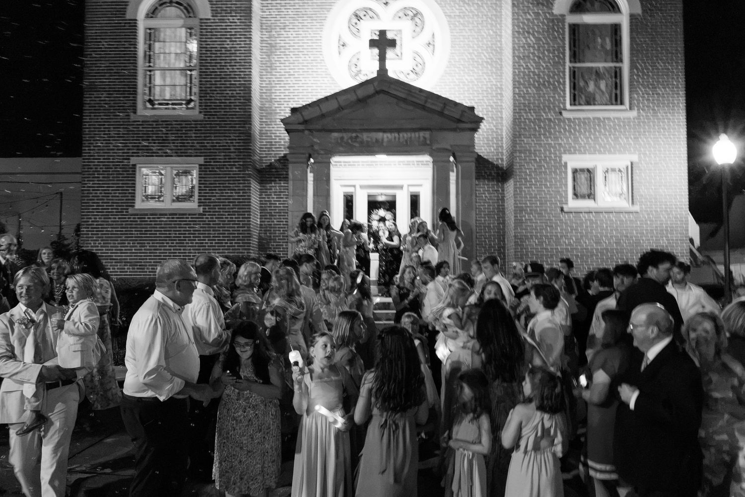 Black and white photo of newlyweds exiting church surrounded by cheering wedding guests at night.