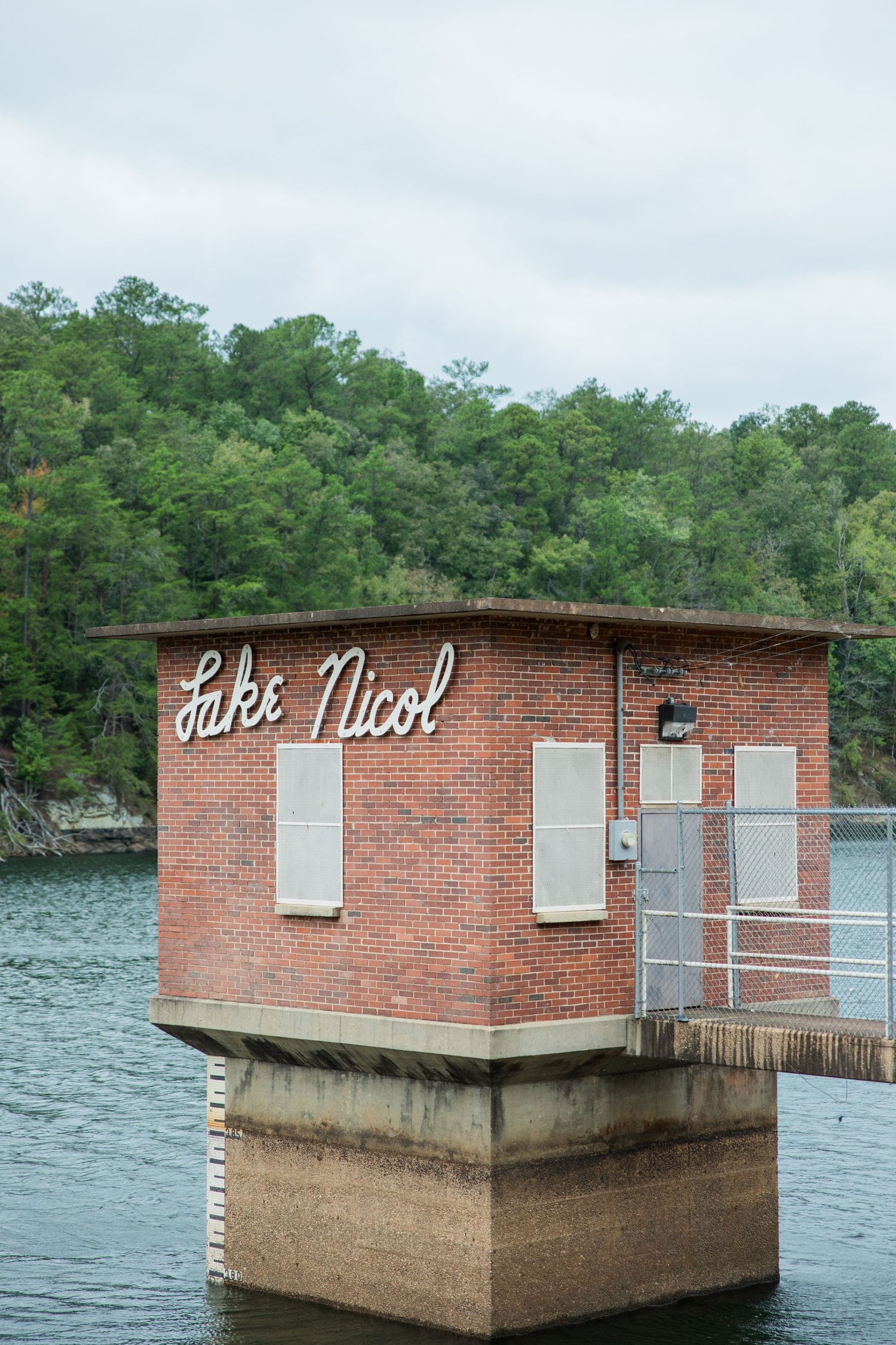 Brick intake tower with a Lake Nicol sign rising from calm water, with a forested green hillside in the background.