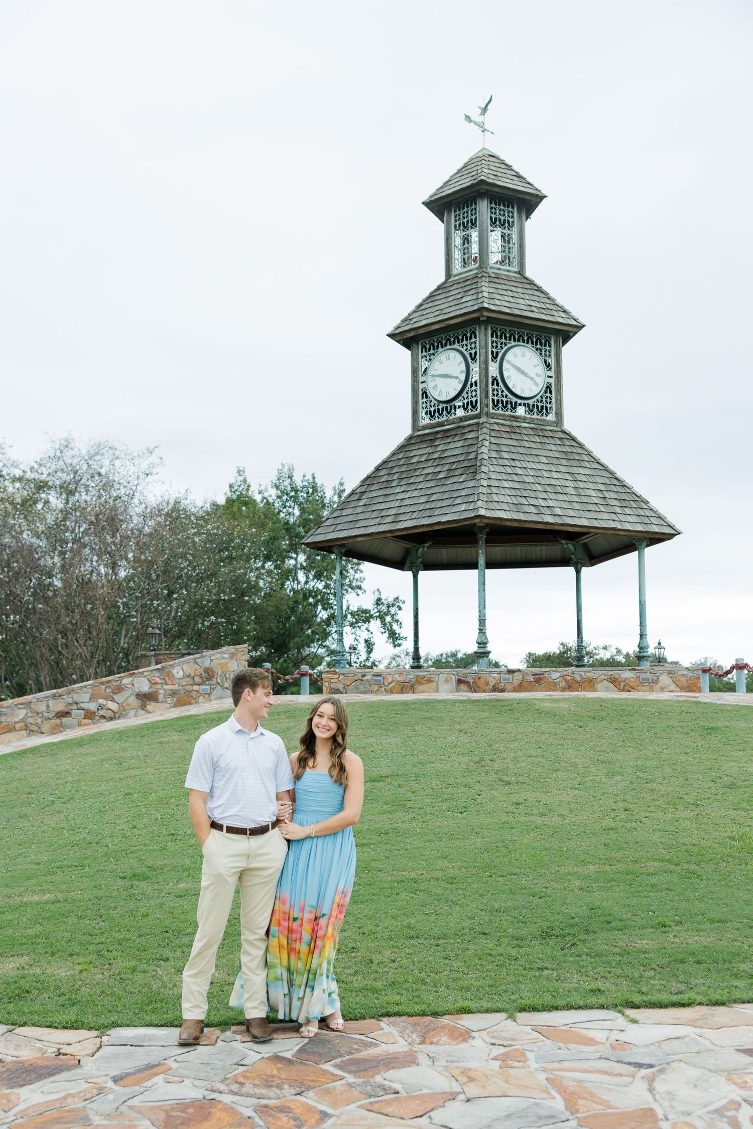 Engaged couple standing hand in hand on a green lawn before a clock tower gazebo with a stone path in the foreground.