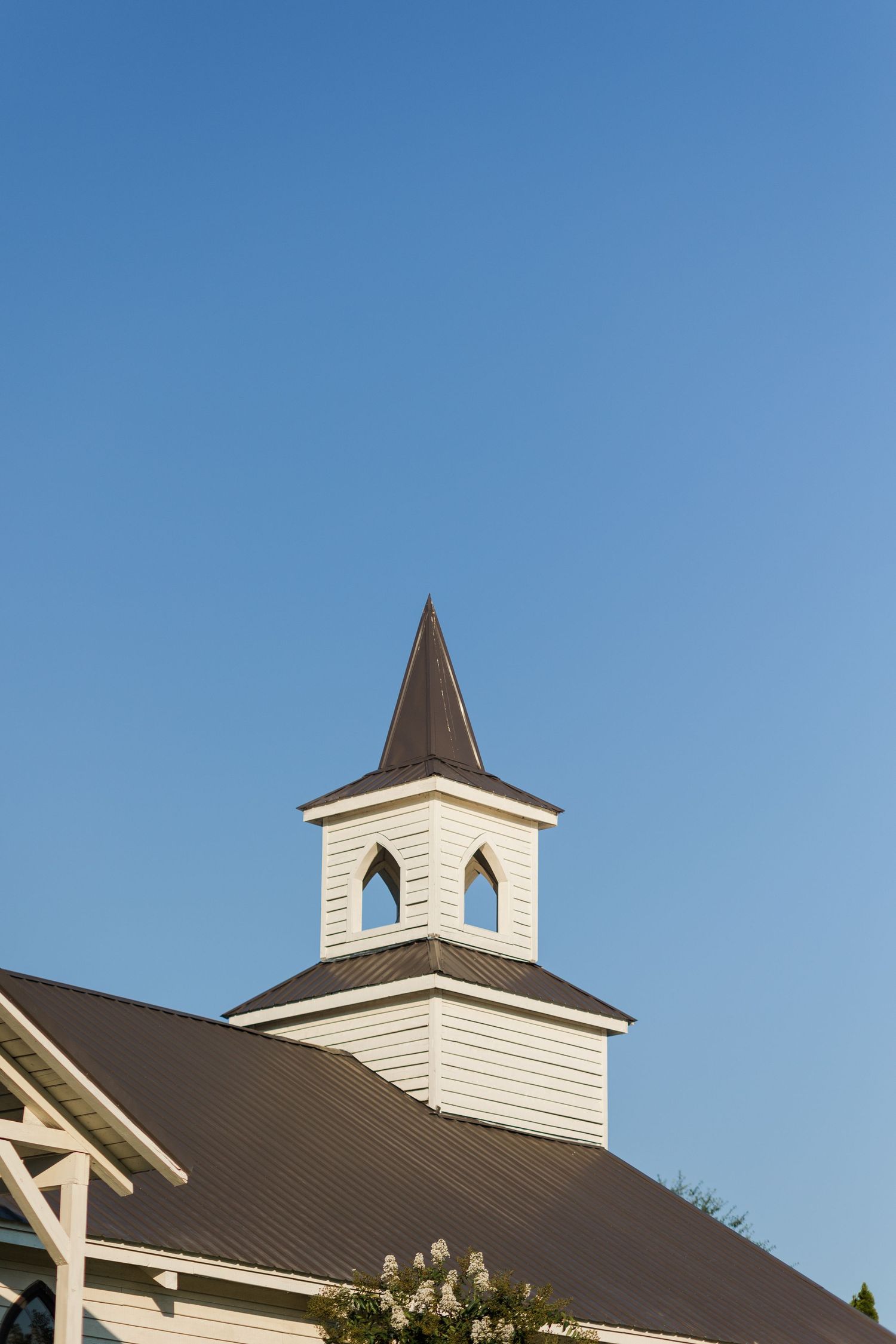 White wooden church steeple with arched windows and dark pointed spire against a clear blue sky.