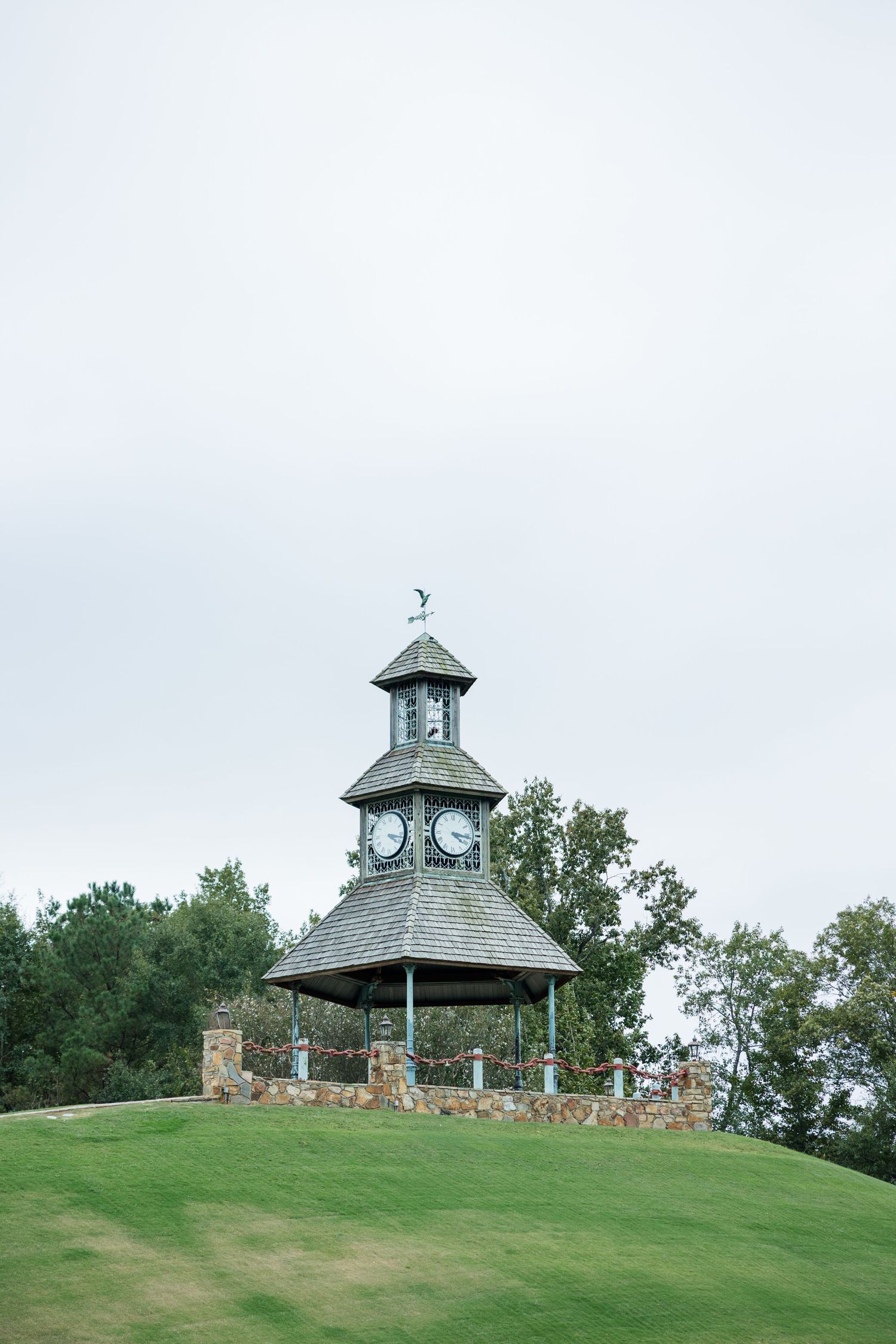 A clock tower gazebo with a gray shingled roof and weathervane sits atop a grassy hill surrounded by trees under an overcast sky.