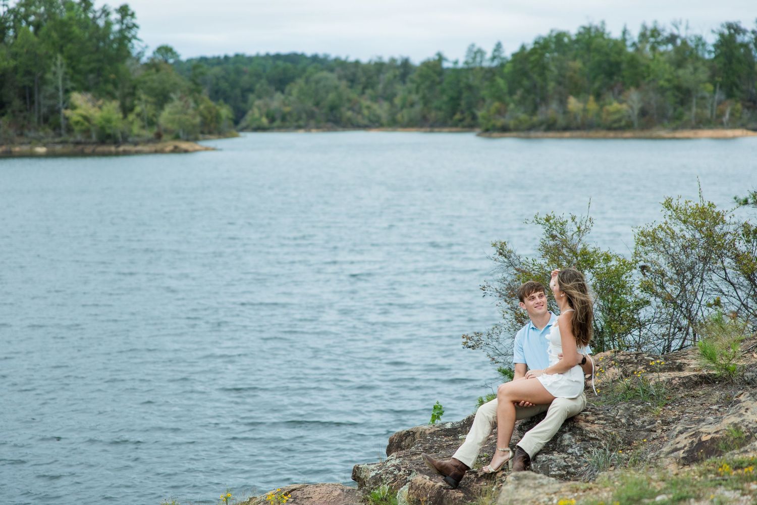 Couple sitting close together on a rocky shore overlooking a calm lake surrounded by forested hills.